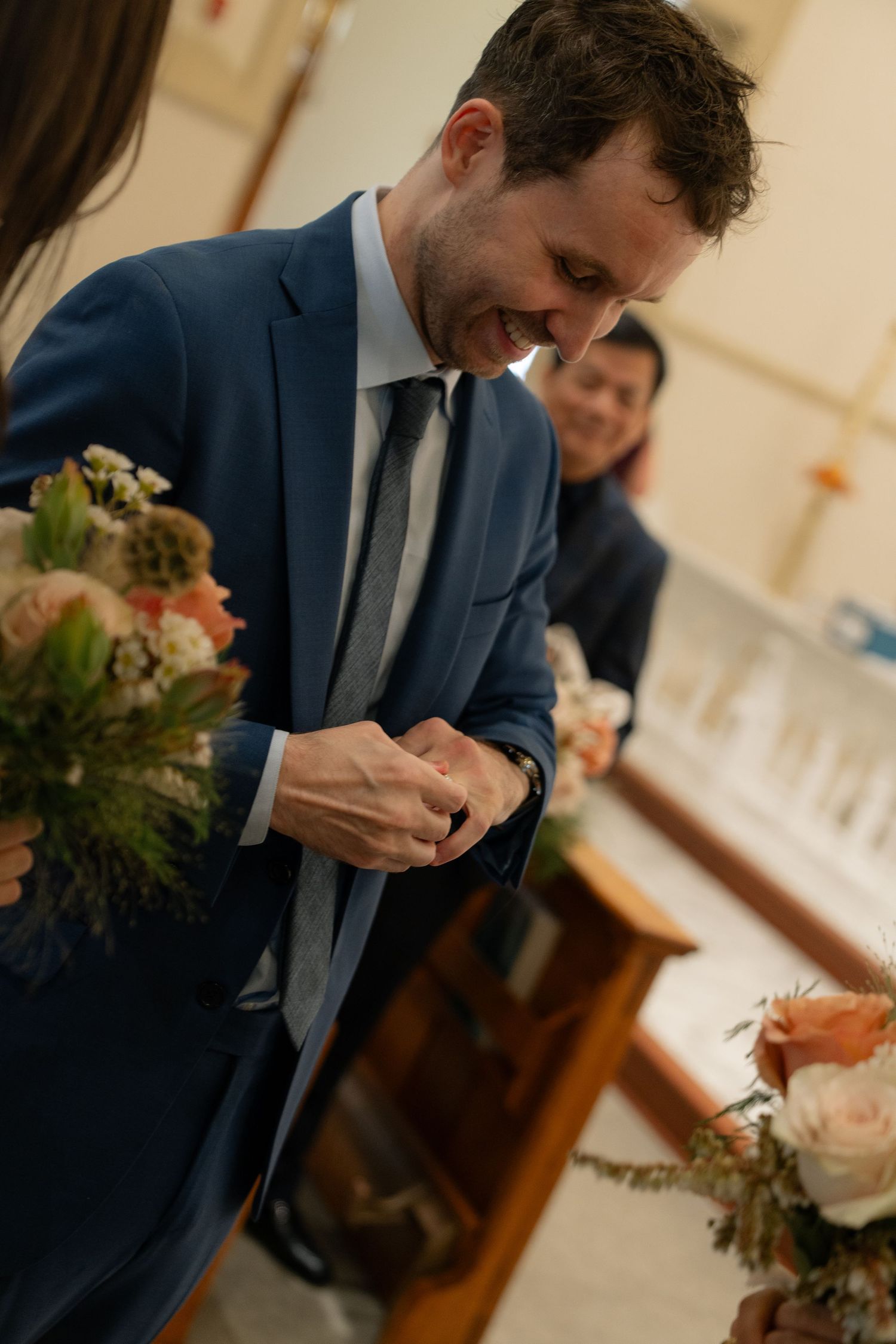 A groom in a navy blue suit smiles while looking down during a church ceremony.