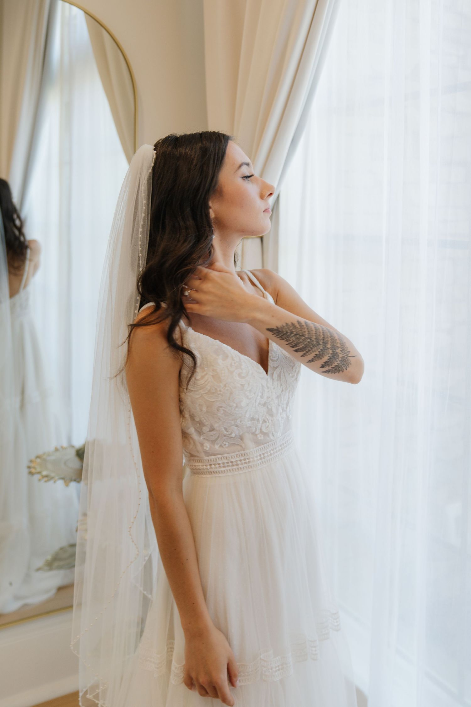 A bride in a white lace wedding dress with a long veil stands by a window in soft natural light.