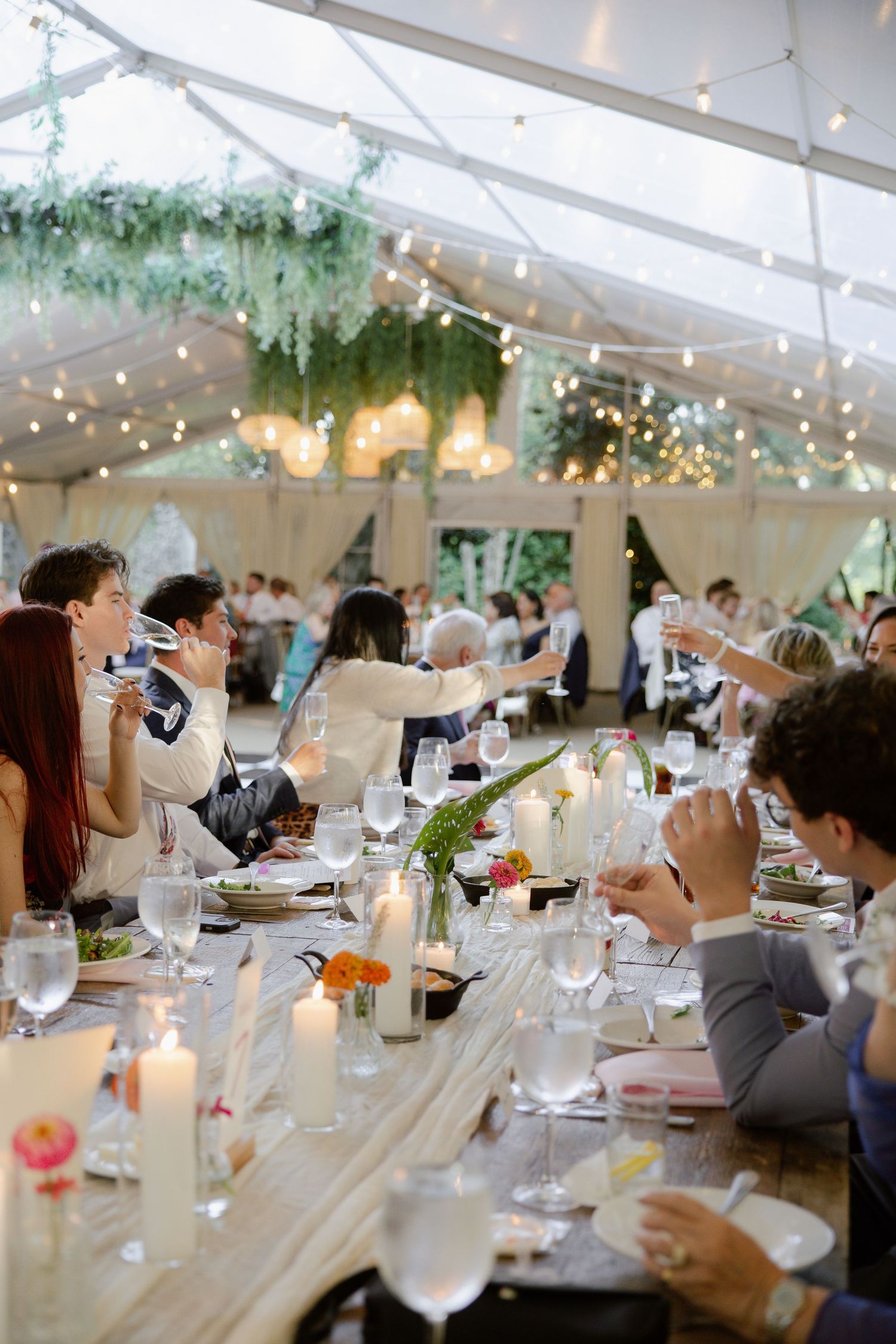 Wedding reception tent decorated with string lights and greenery over elegant table settings.