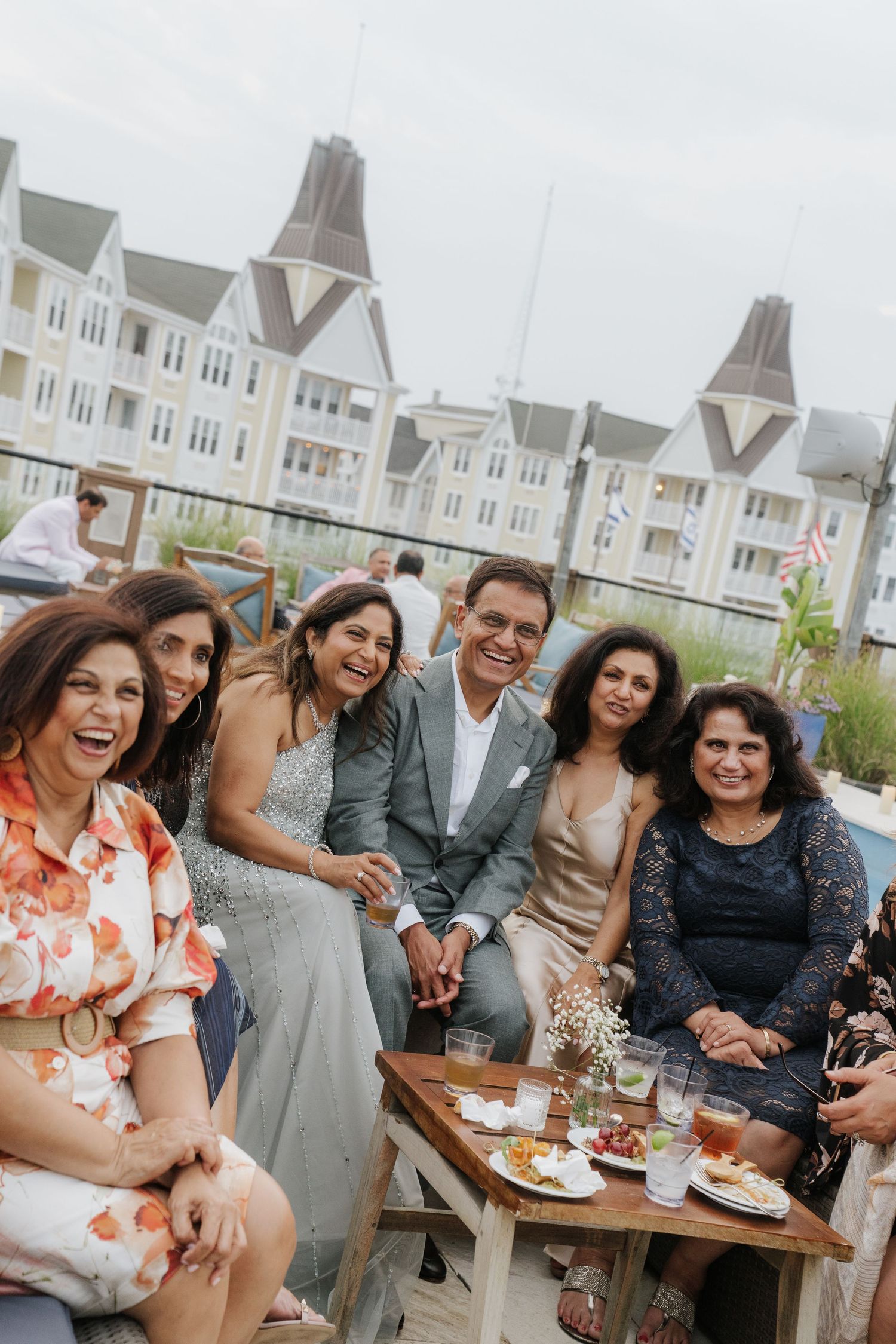 Wedding party enjoying drinks and conversation at outdoor reception with coastal buildings in background.