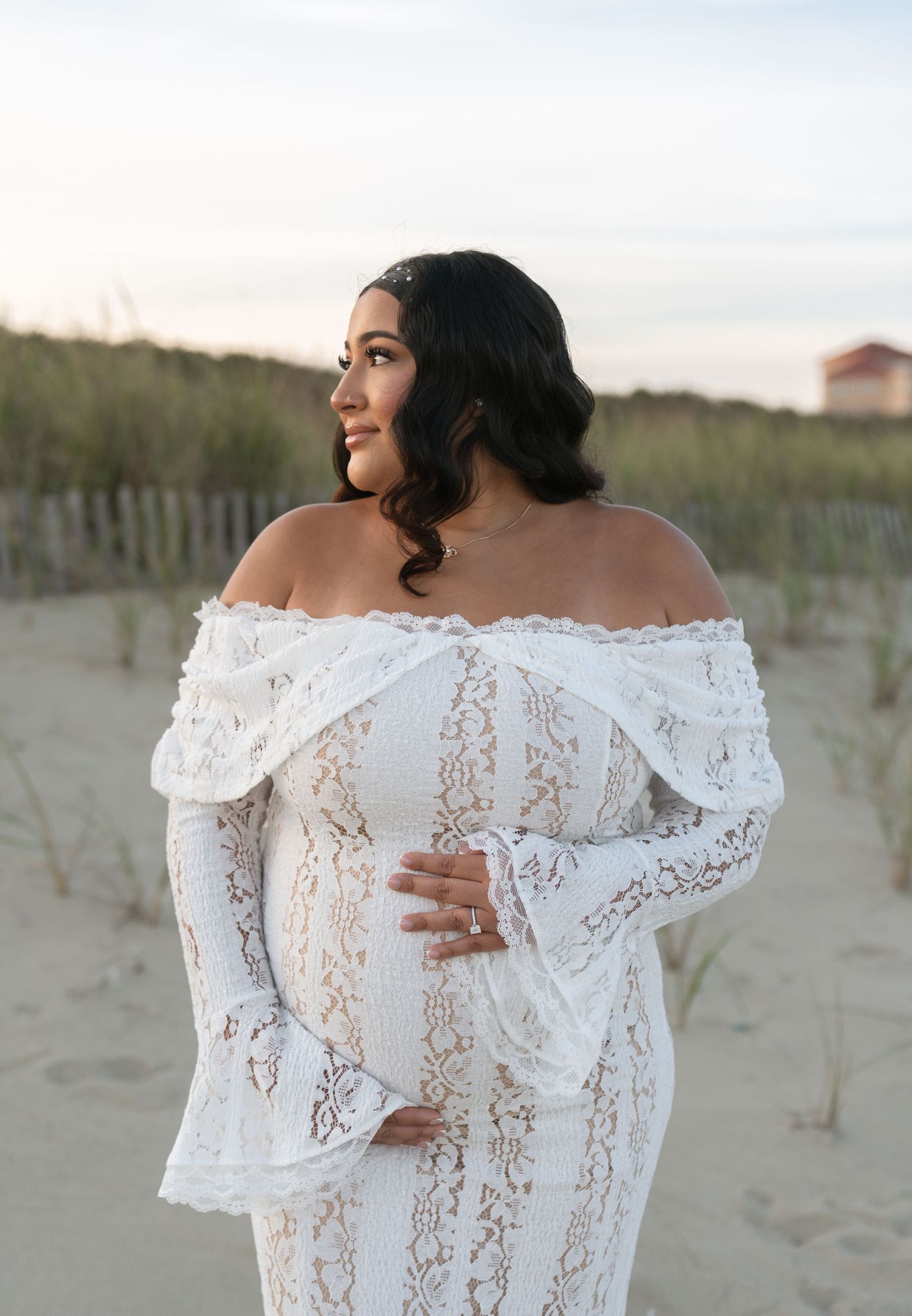 Pregnant woman in white off-shoulder lace gown poses on sandy beach at sunset, cradling her baby bump.