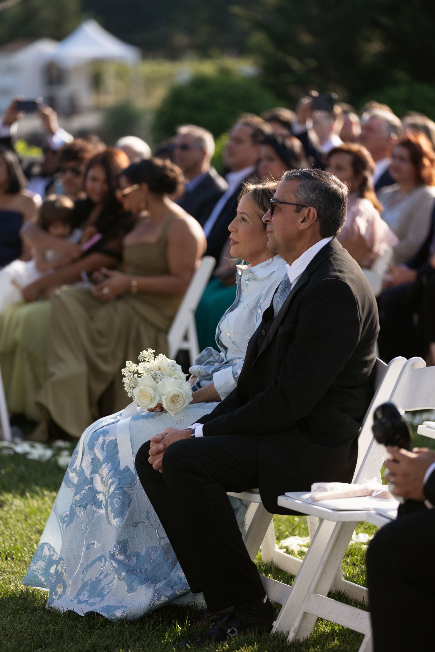 Couple in formal attire sits in white chairs at an outdoor wedding ceremony with guests.