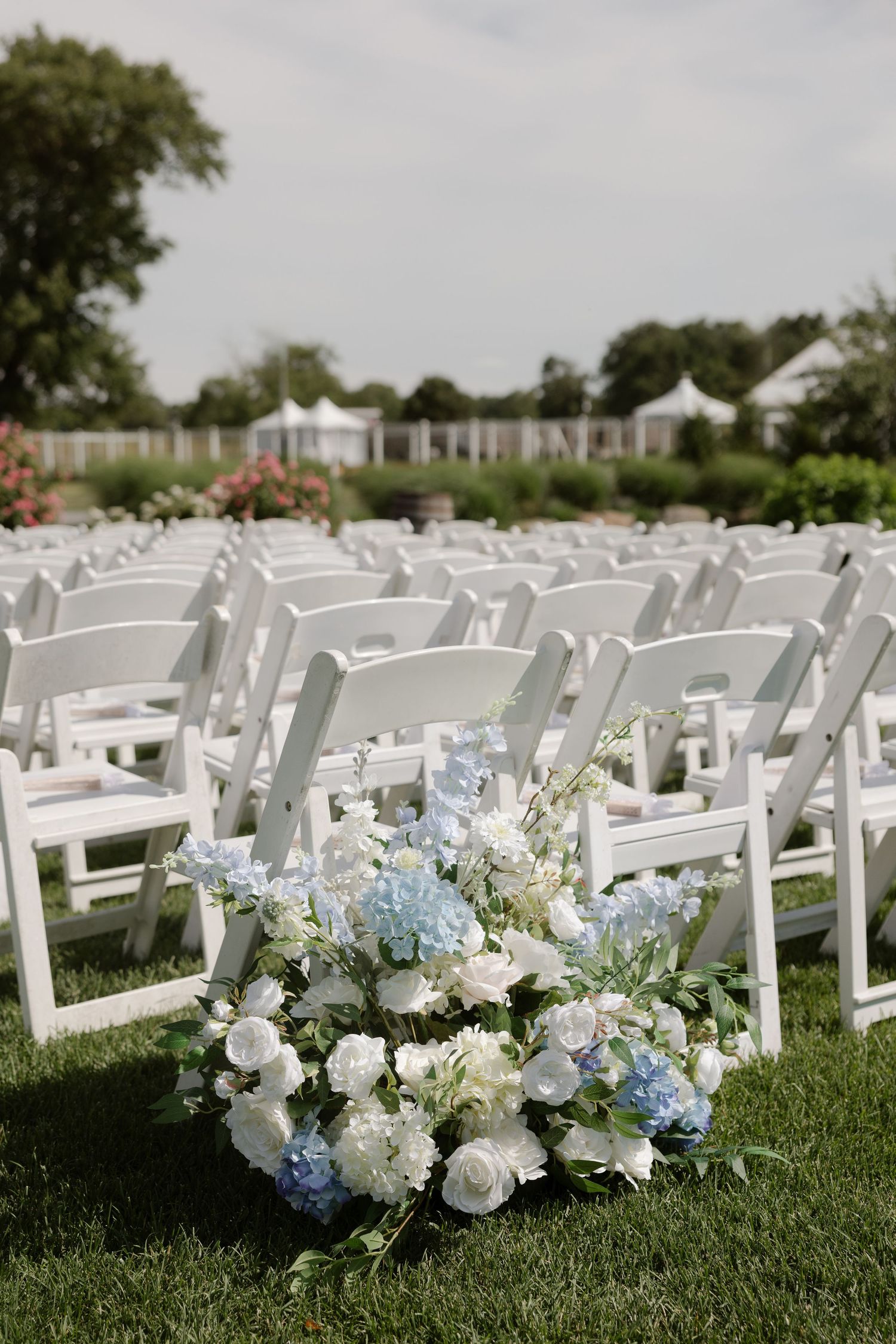 White floral arrangement with blue hydrangeas beside rows of white folding chairs at outdoor wedding ceremony.