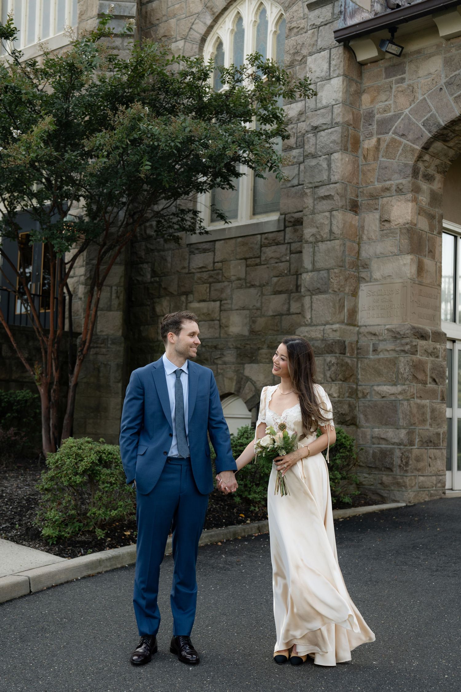 A couple holds hands outside a stone church building with gothic architecture and landscaping.
