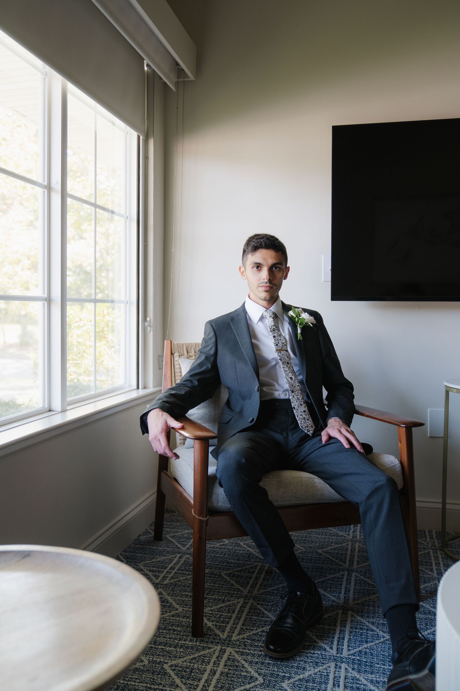 A person in a gray suit sits in a chair by a window in a minimalist room with patterned carpet.