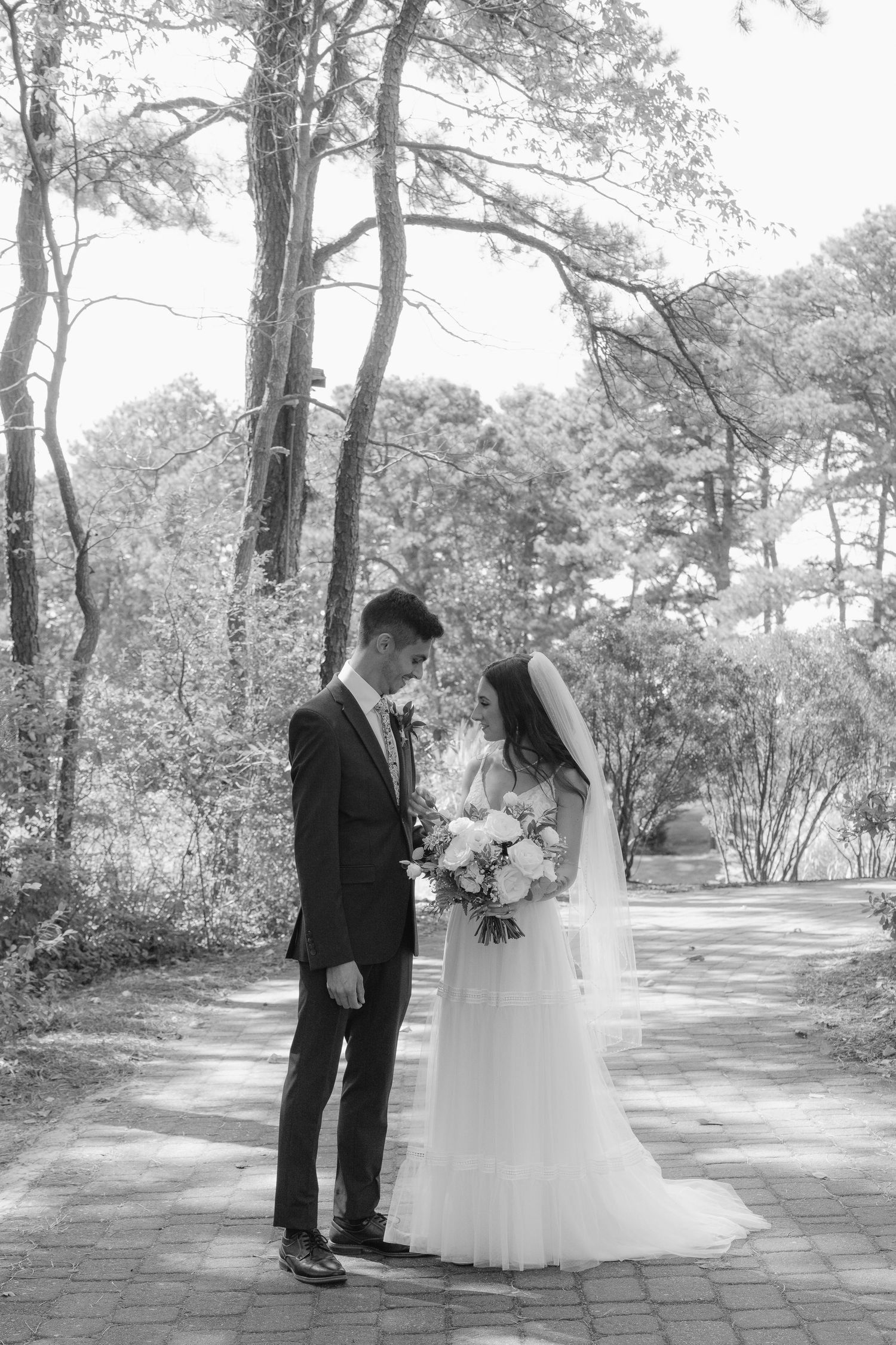 A romantic wedding couple shares tender moments along a tree-lined garden path in black and white photography.