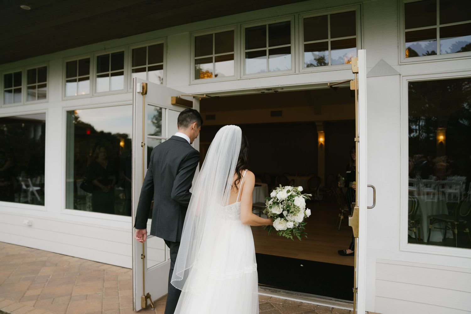 A wedding couple walks along a covered outdoor porch decorated with string lights and white columns.