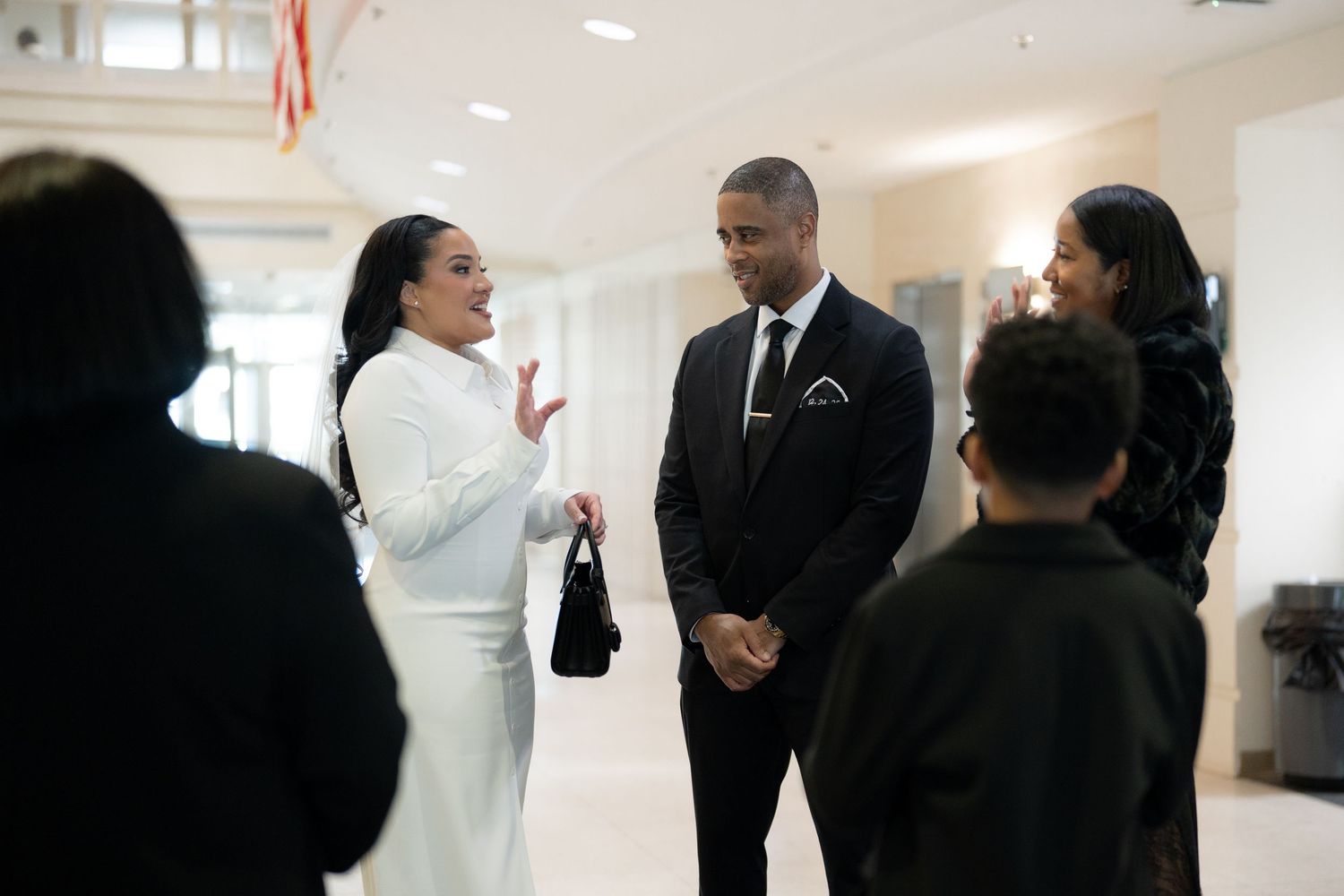 People in formal attire interact in a bright indoor hallway with white walls and carpeted flooring.