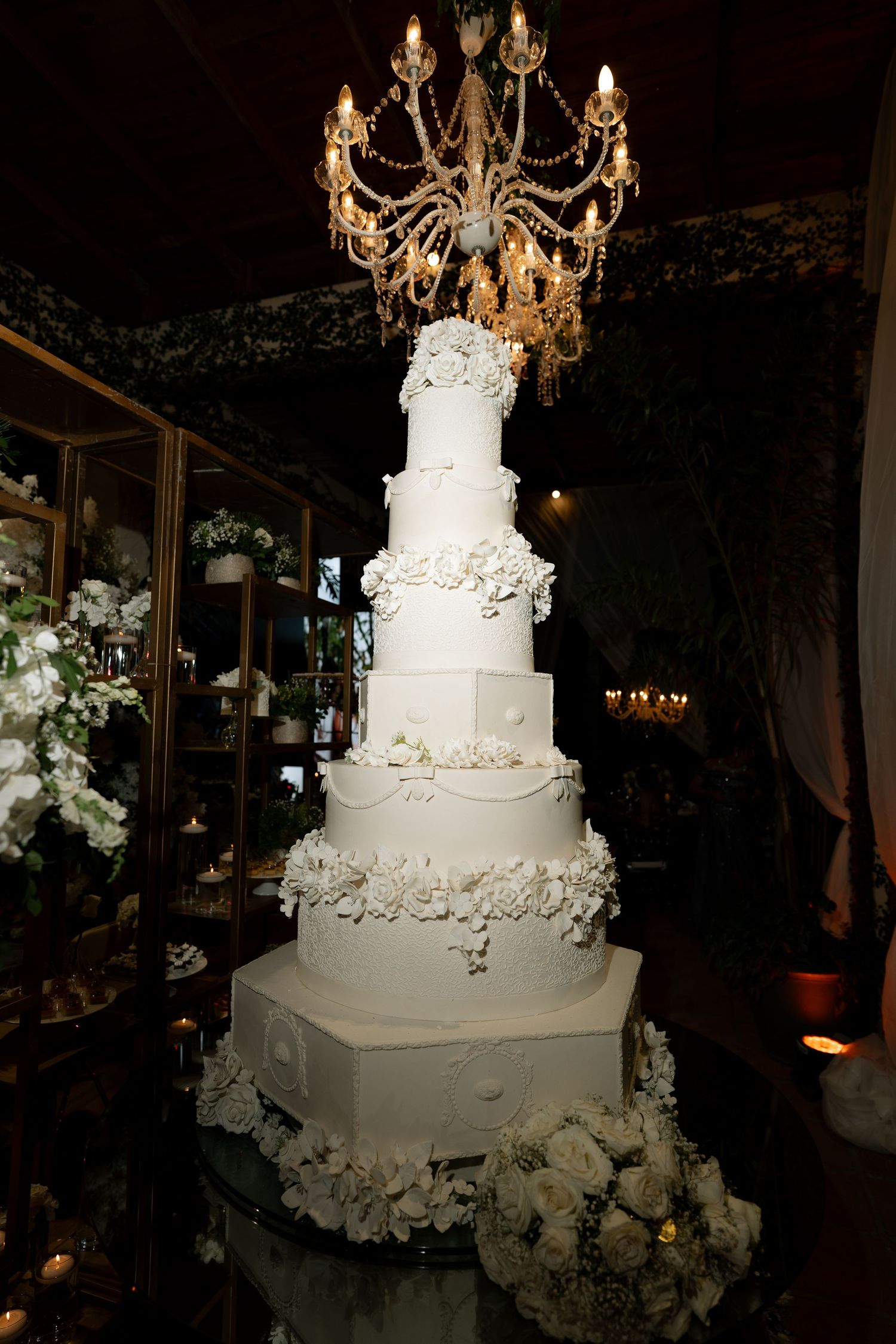 Elegant seven-tier white wedding cake adorned with floral details beneath a crystal chandelier in a luxurious venue.