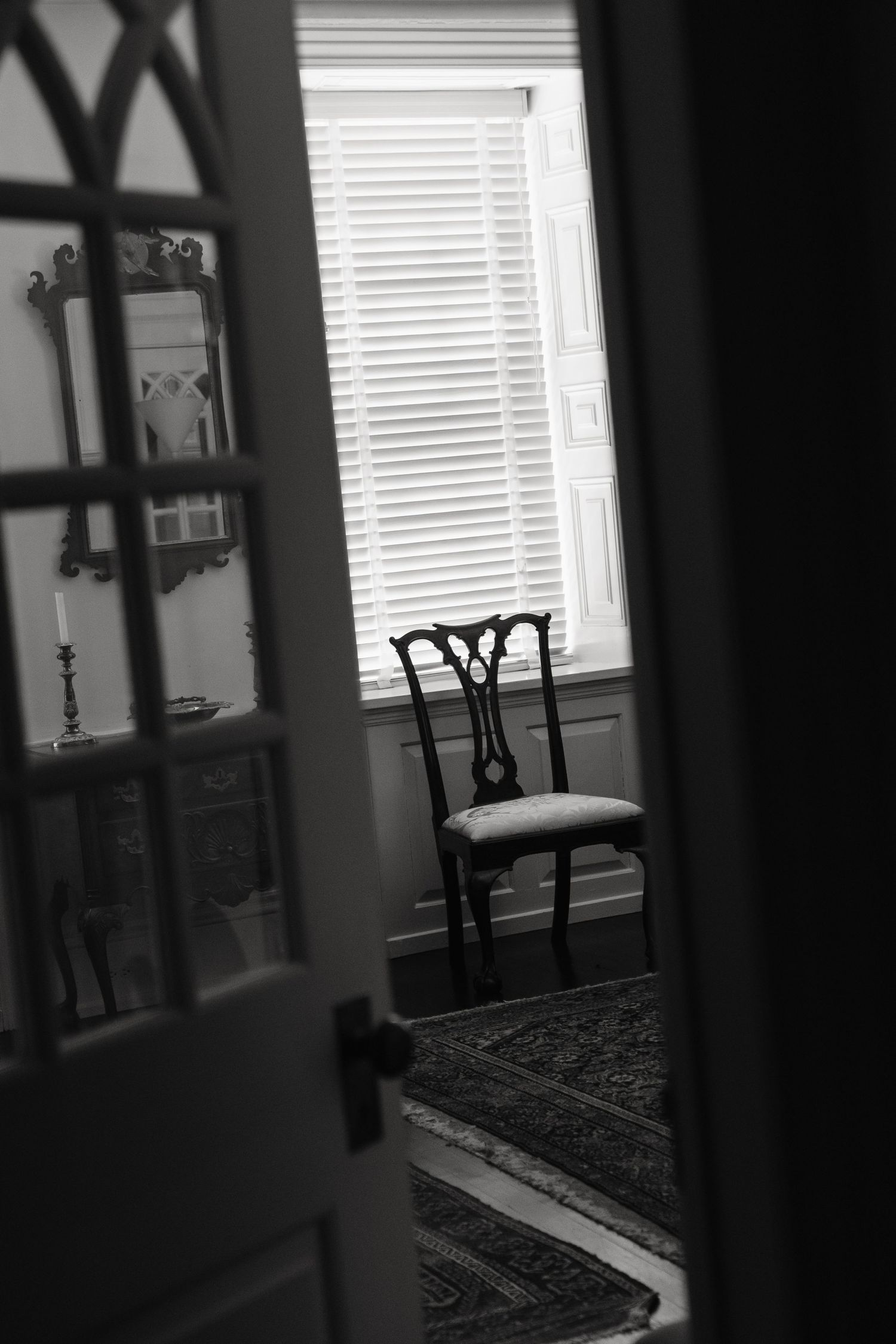 Black and white view through doorway showing empty chair by window with venetian blinds and patterned rug on floor.