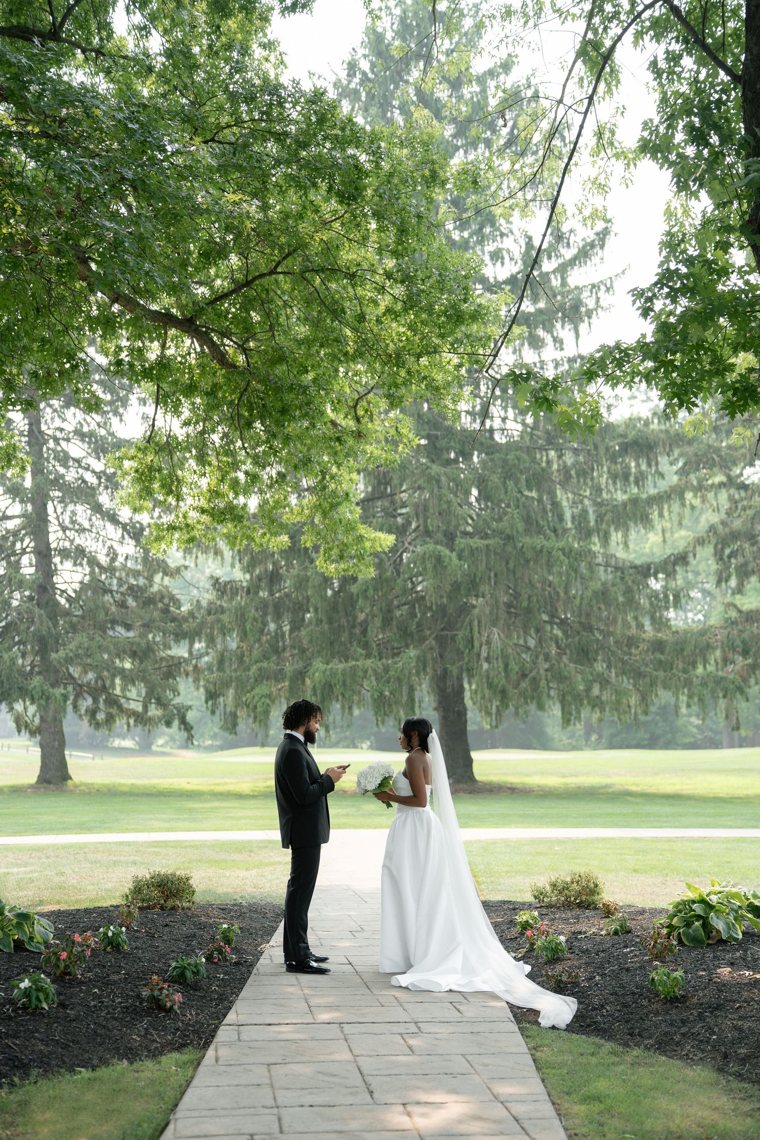 Bride and groom exchange vows on a stone path surrounded by lush green trees in an outdoor wedding ceremony.