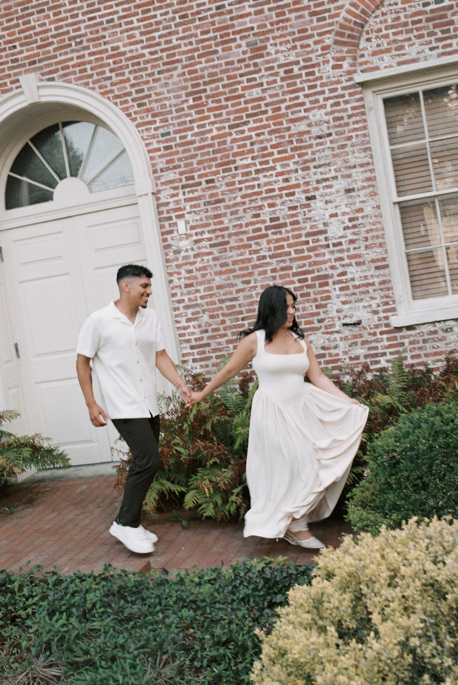 Couple holding hands walking in front of a brick building, woman in white dress, man in white shirt and dark pants.