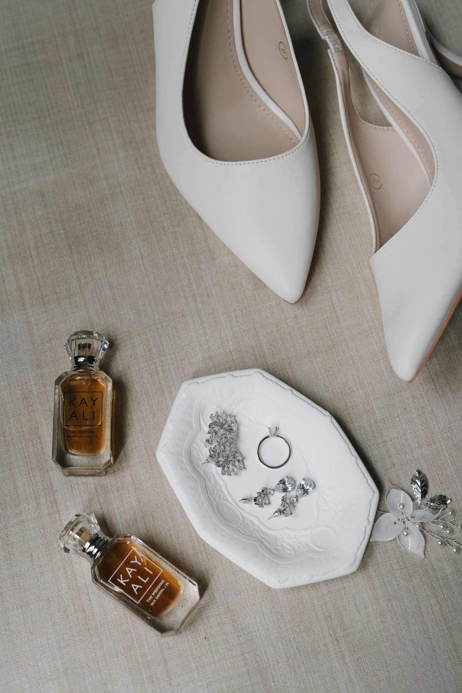 Bridal accessories flat lay featuring white heels, diamond ring, earrings, and amber perfume bottles on linen.