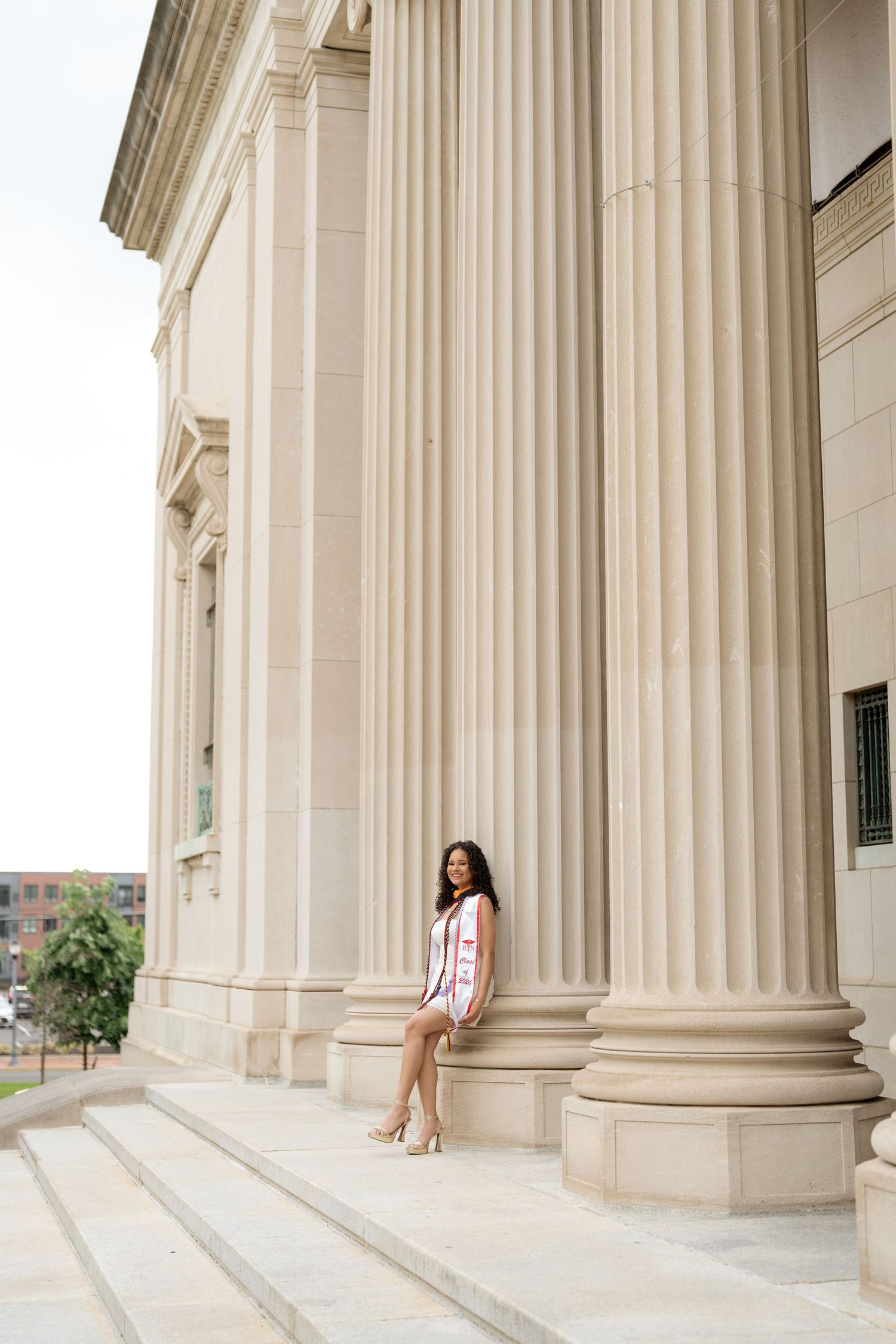 Young woman in pink dress poses on steps of neoclassical building with tall fluted columns.