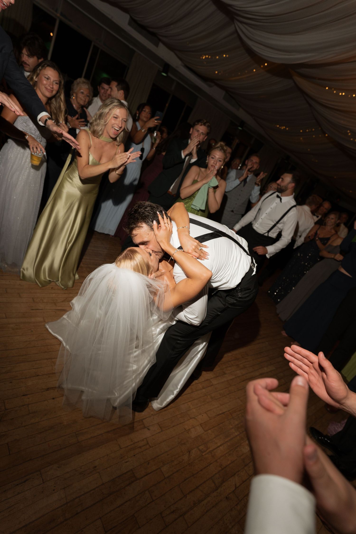 Groom dips bride for a romantic kiss on the dance floor surrounded by cheering wedding guests.