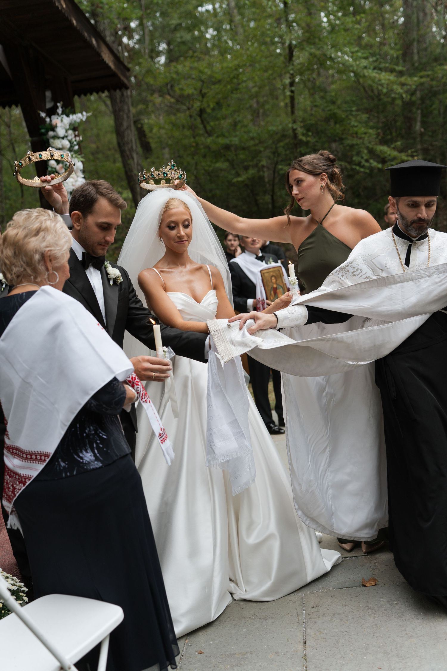 Orthodox wedding ceremony with priest, bride, groom, and attendants holding crowns and candles outdoors in a forest.
