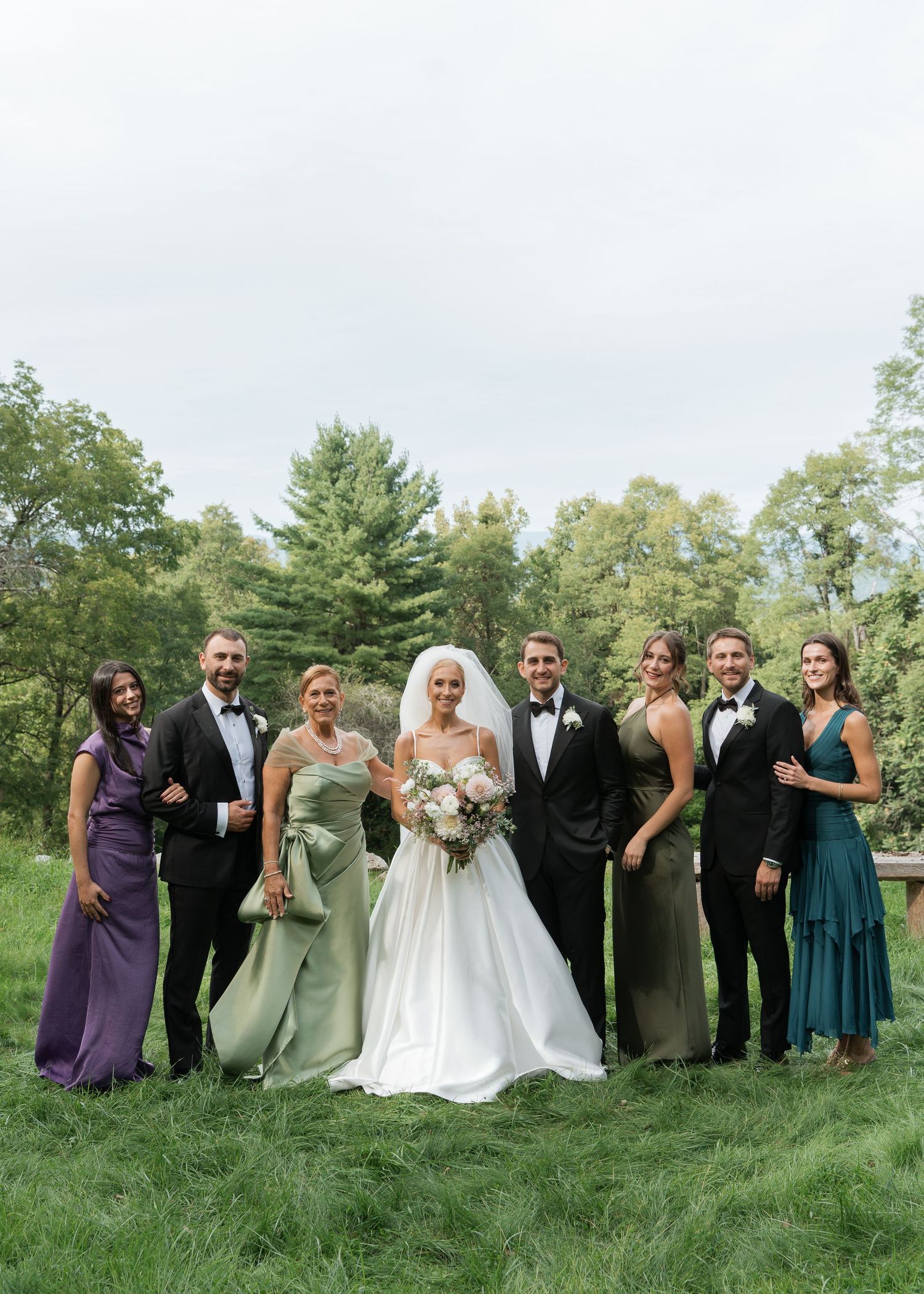 Wedding party poses outdoors in elegant attire, bride in white gown with veil, bridesmaids in green and purple dresses.