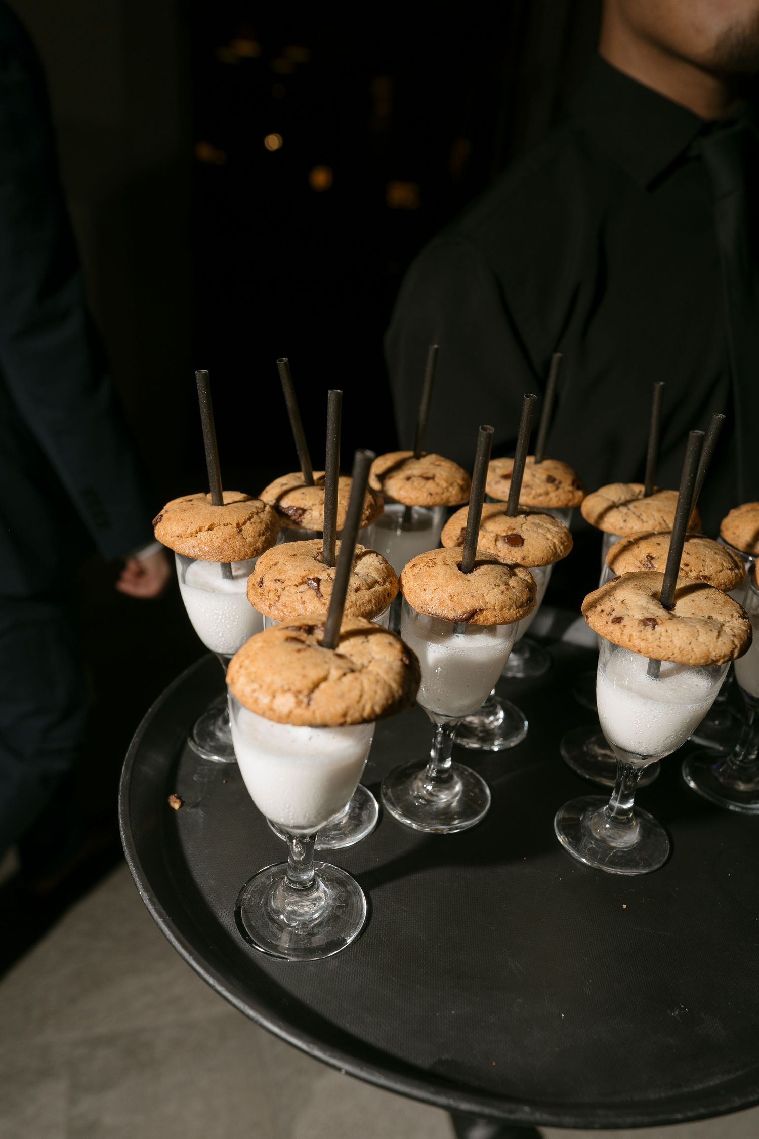 Server holding tray of milk and cookie dessert shooters topped with chocolate chip cookies and straws.