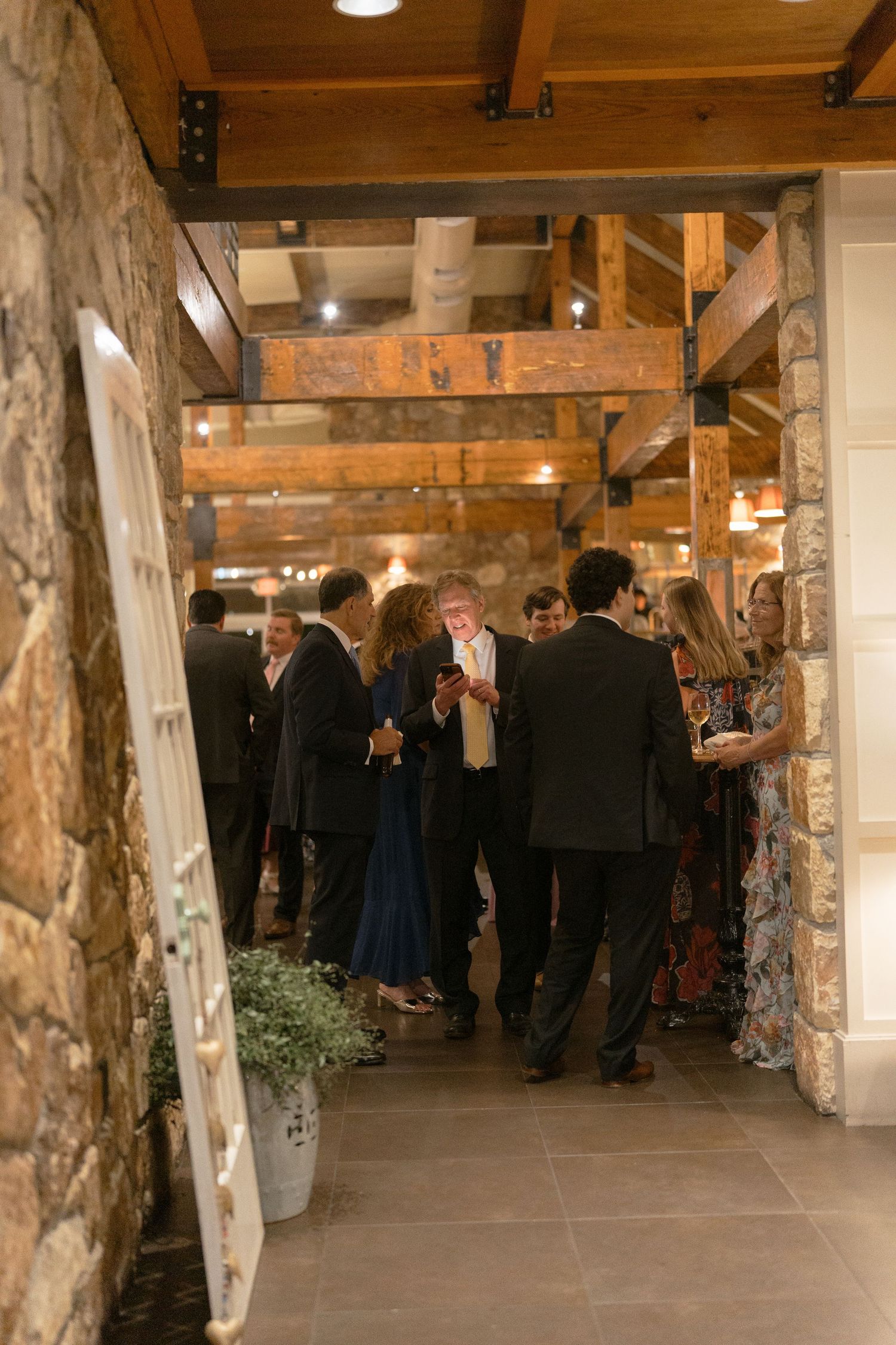 Wedding guests mingle in rustic stone and wood beam venue during cocktail hour reception gathering.