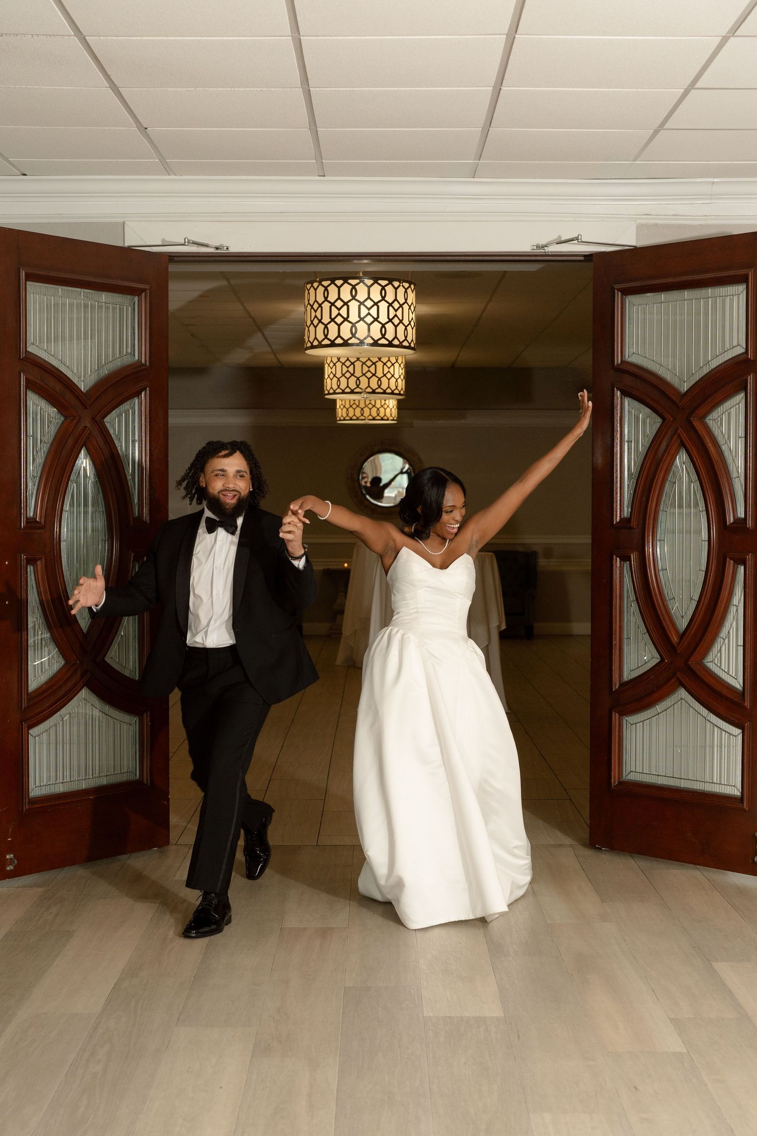 Jubilant newlyweds make grand entrance through elegant wooden doors at wedding reception venue.