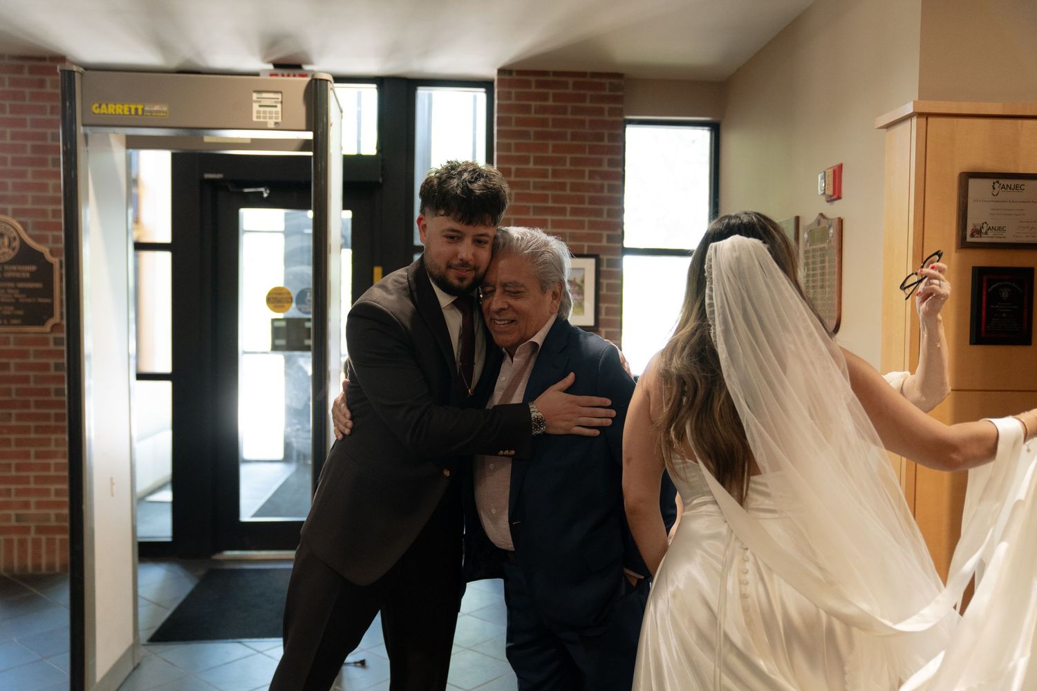 Bride and groom embracing an elderly guest in a wedding venue hallway, with the bride in a white gown and veil.