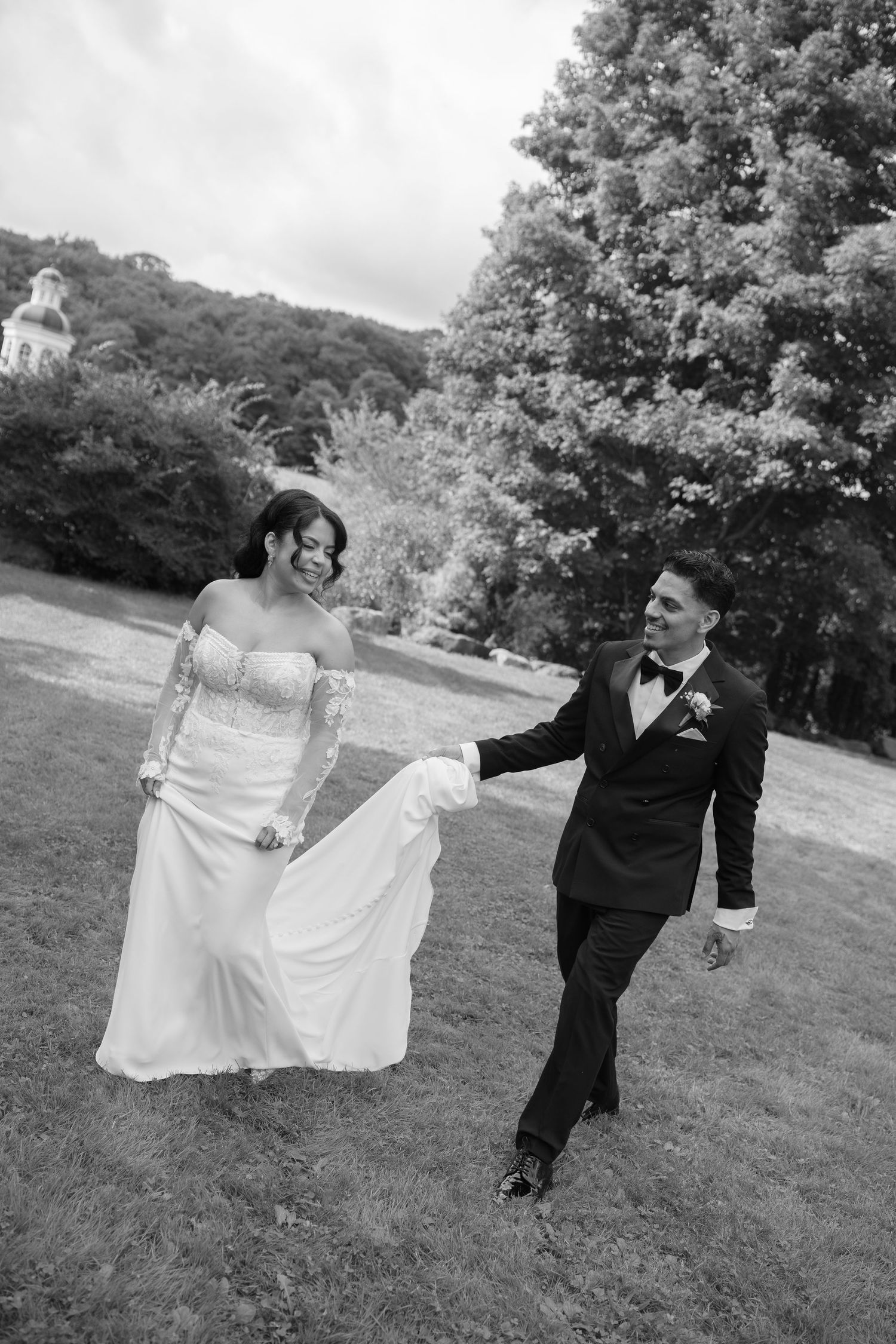 Black and white photo of a bride and groom walking hand in hand across a lush green lawn on their wedding day.