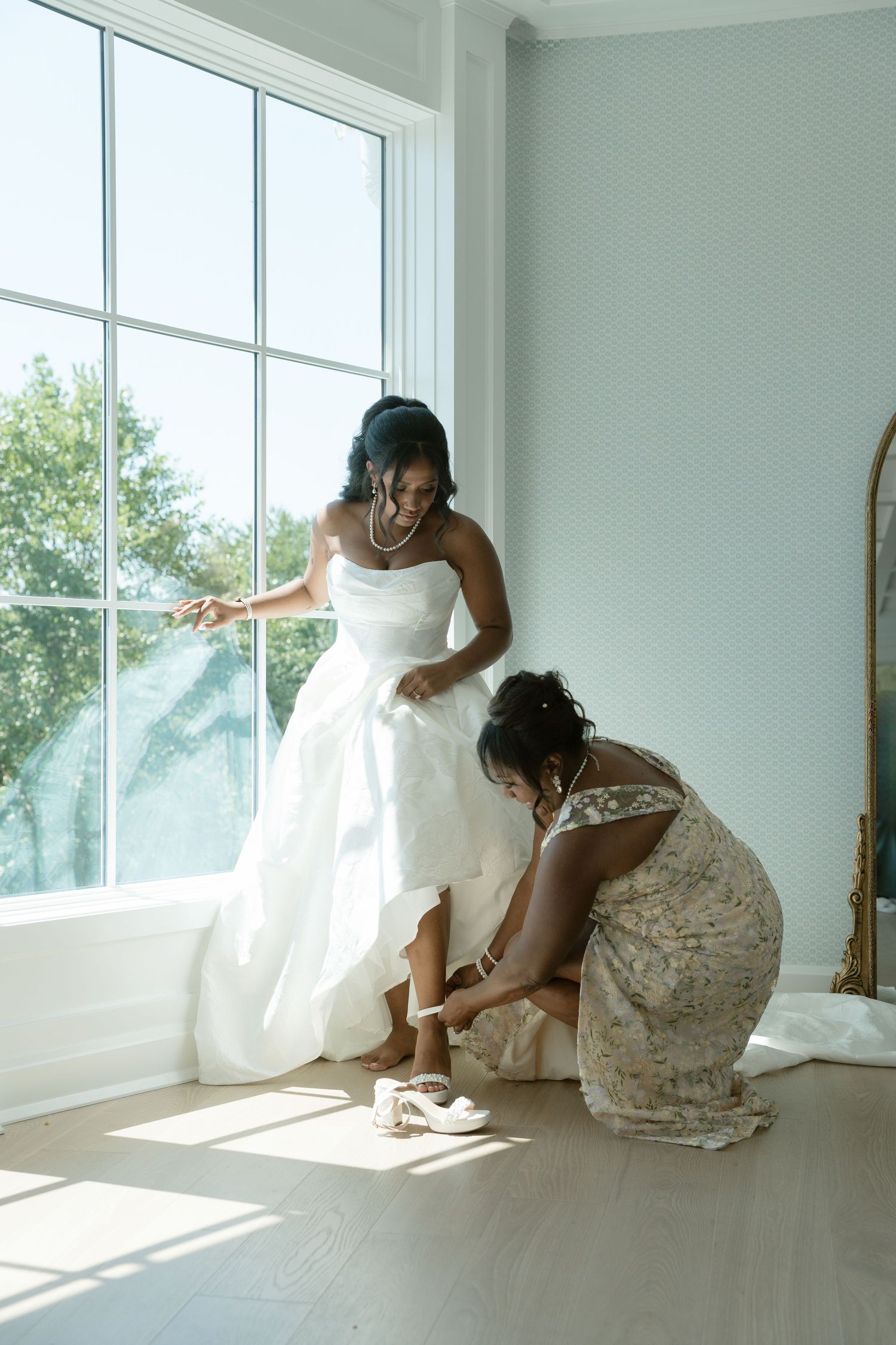 Bride in white gown gets shoes fastened by bridesmaid near sunlit window in elegant bridal suite.