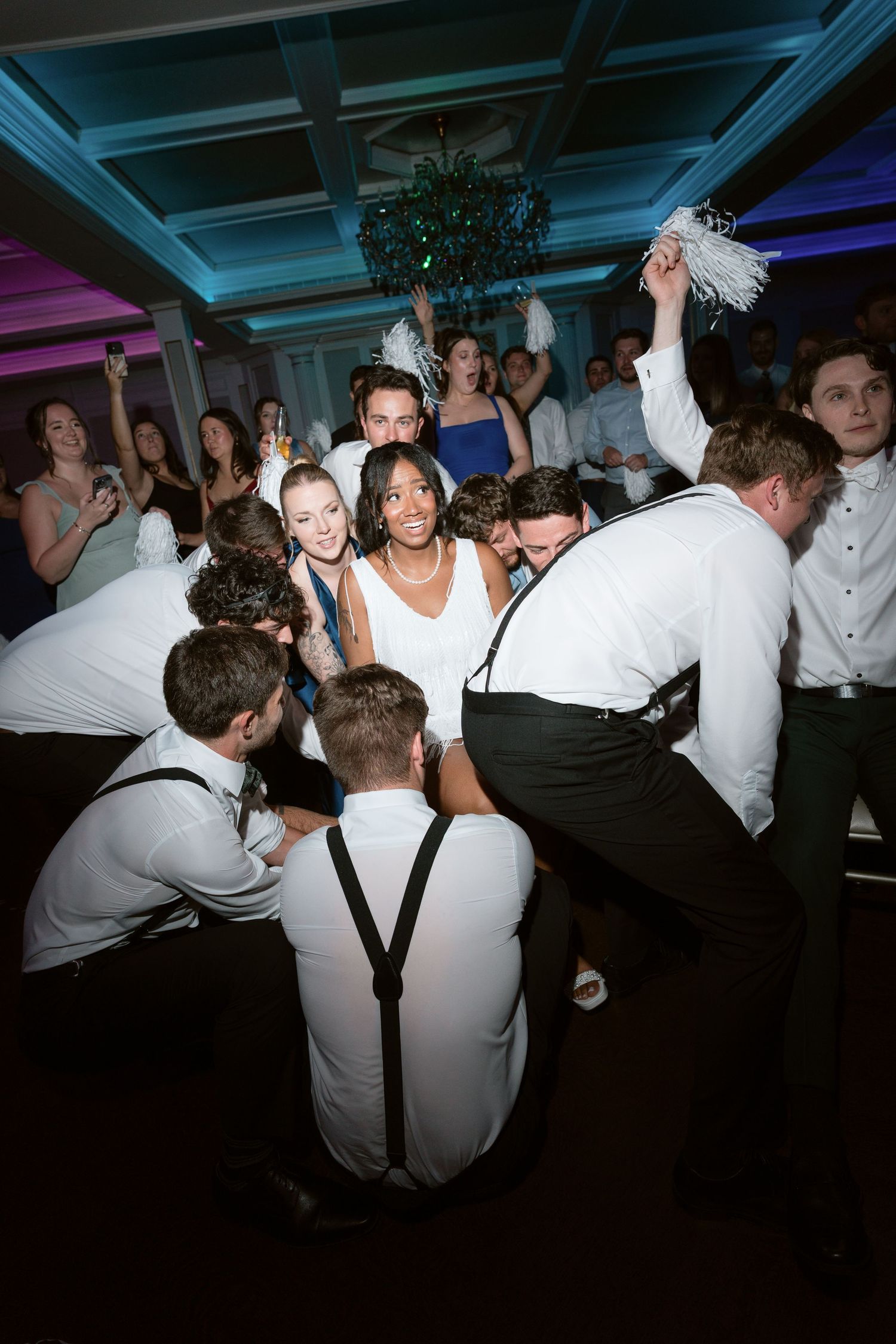Bride surrounded by groomsmen in white shirts during lively wedding reception dance floor celebration.