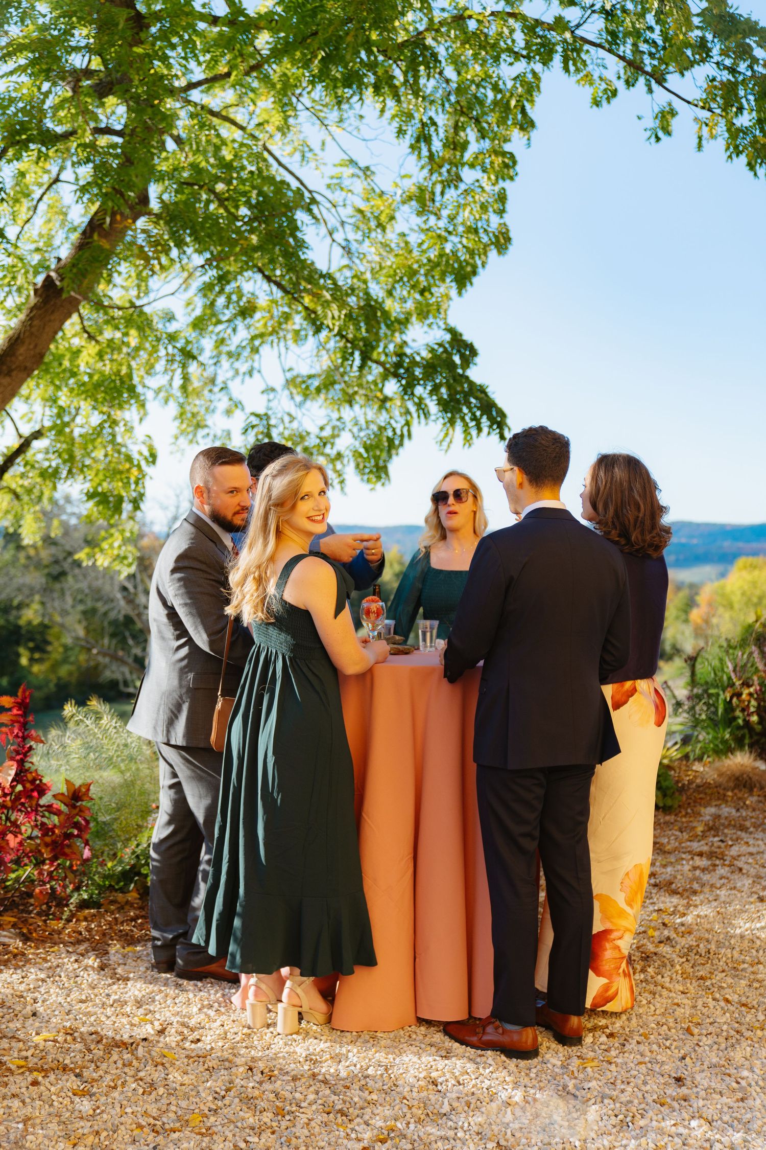 Wedding party in green and peach dresses posing together outdoors under leafy trees.