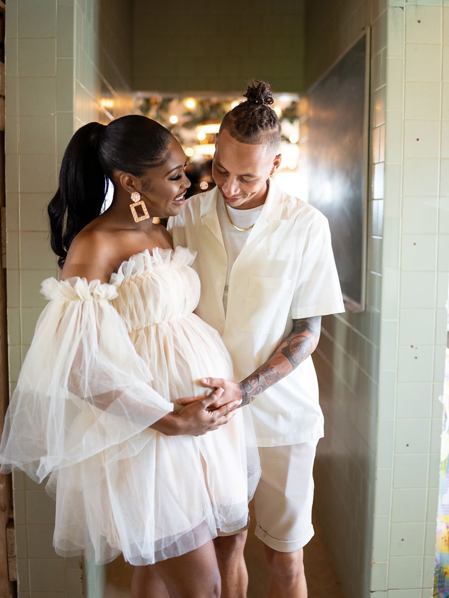 A couple shares a tender moment in matching white outfits against a light-filled hallway.