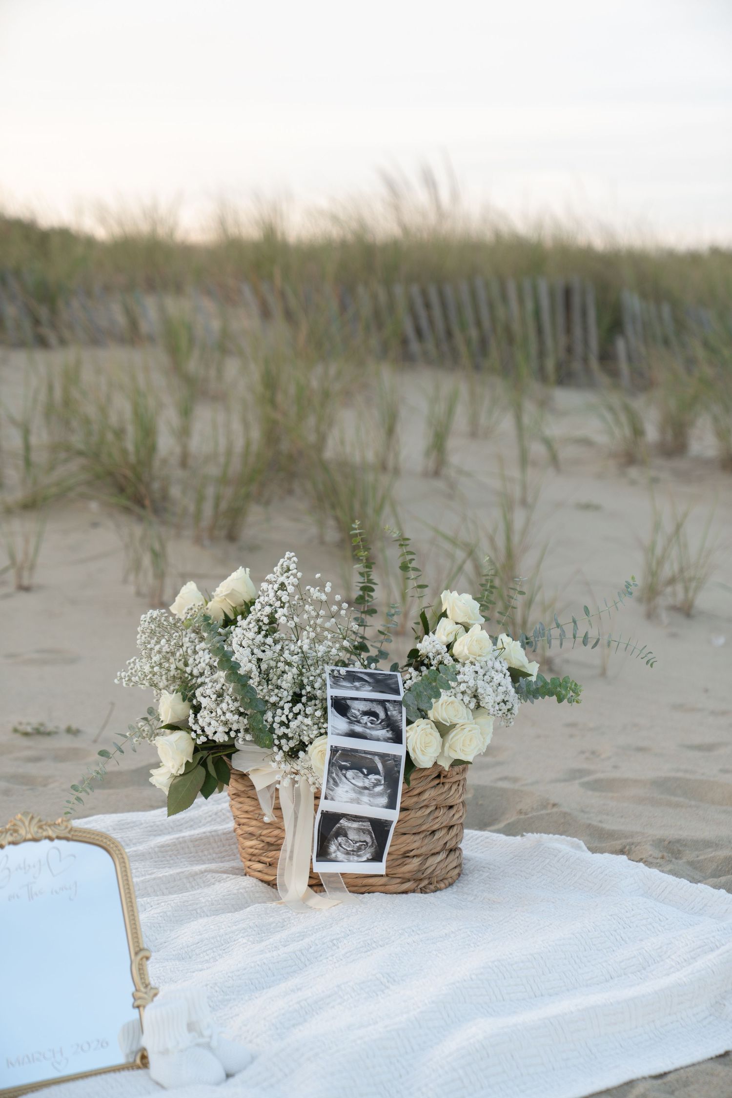 A beach scene with ultrasound photos and white flowers arranged on a blanket against sand dunes.