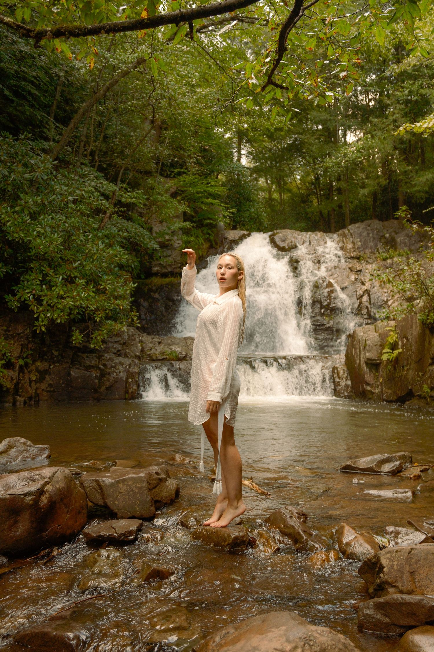 Figure in flowing white dress poses gracefully against scenic waterfall backdrop in nature.