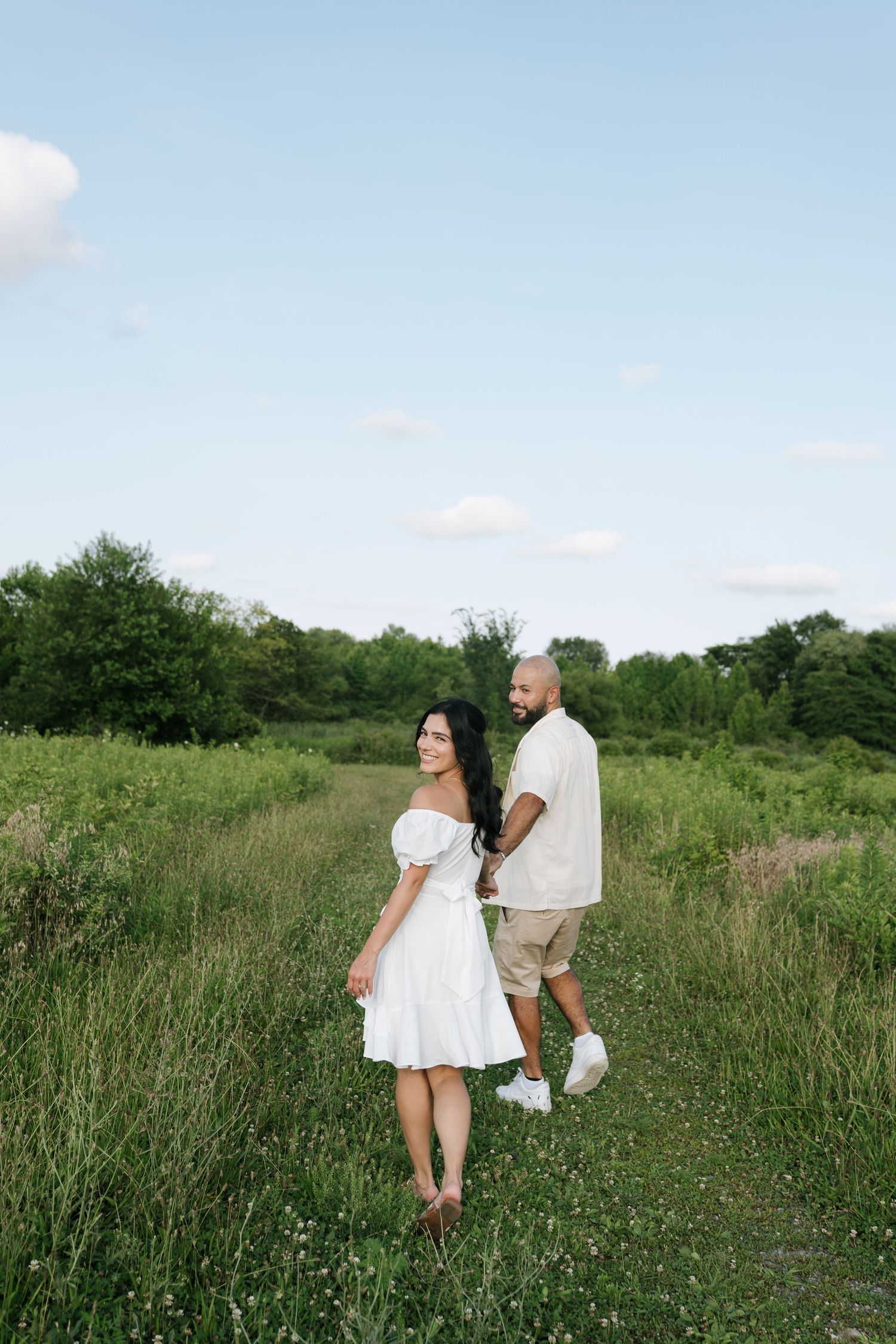 A couple walks through a sunny wildflower meadow in wedding attire.