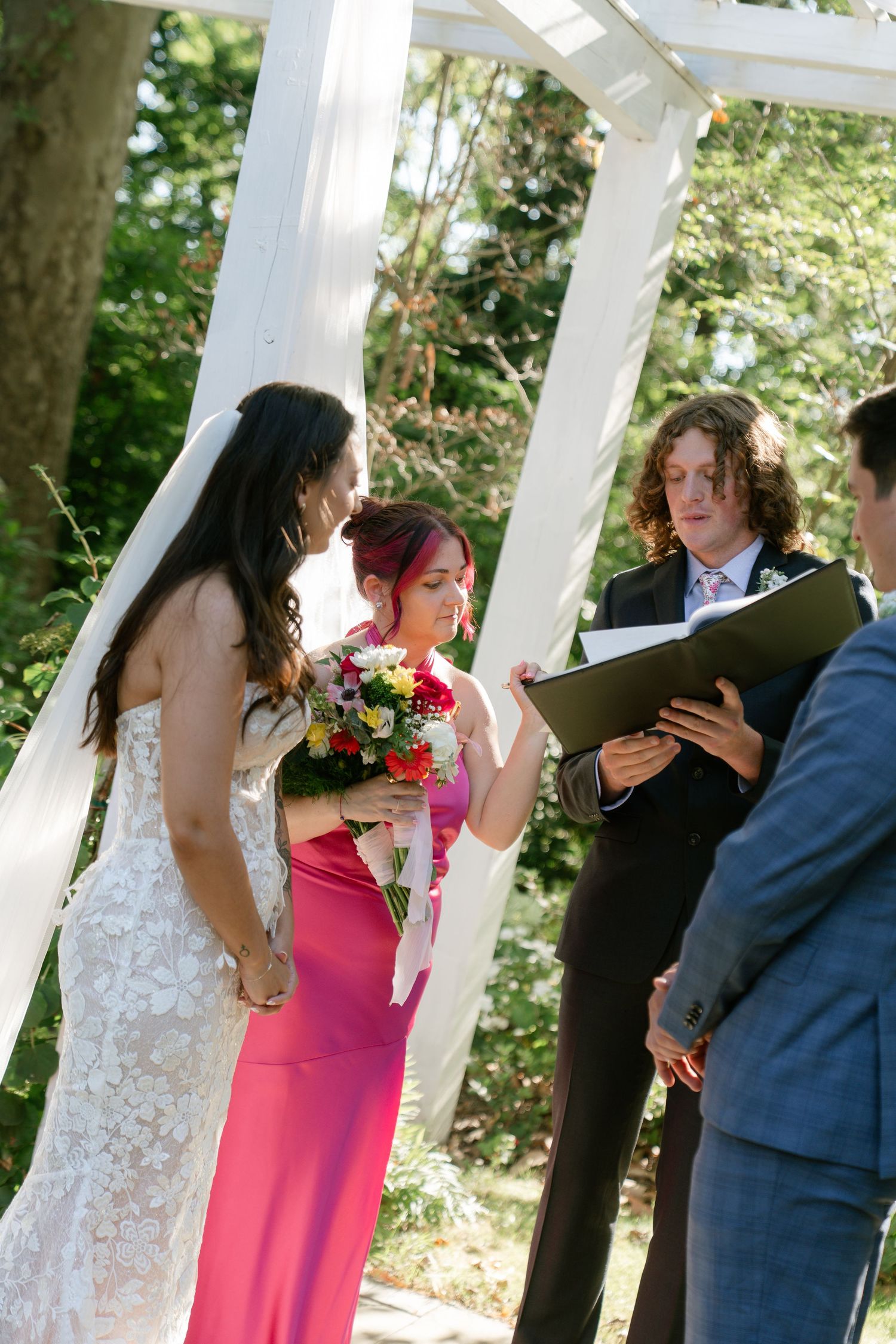 Wedding party members stand under a white gazebo during an outdoor ceremony.