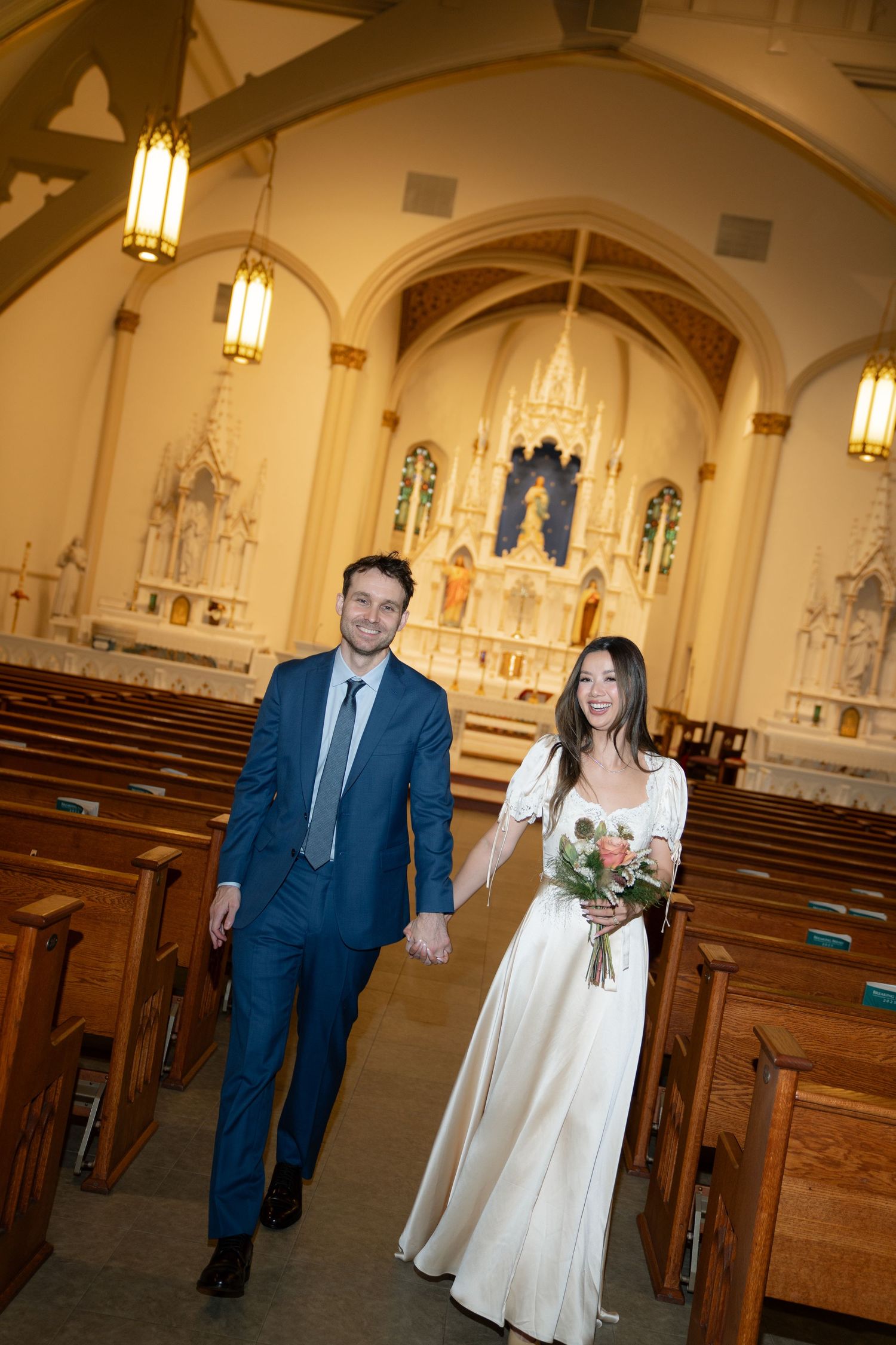 A couple walks down the aisle of an ornate church with vaulted ceilings and stained glass windows.