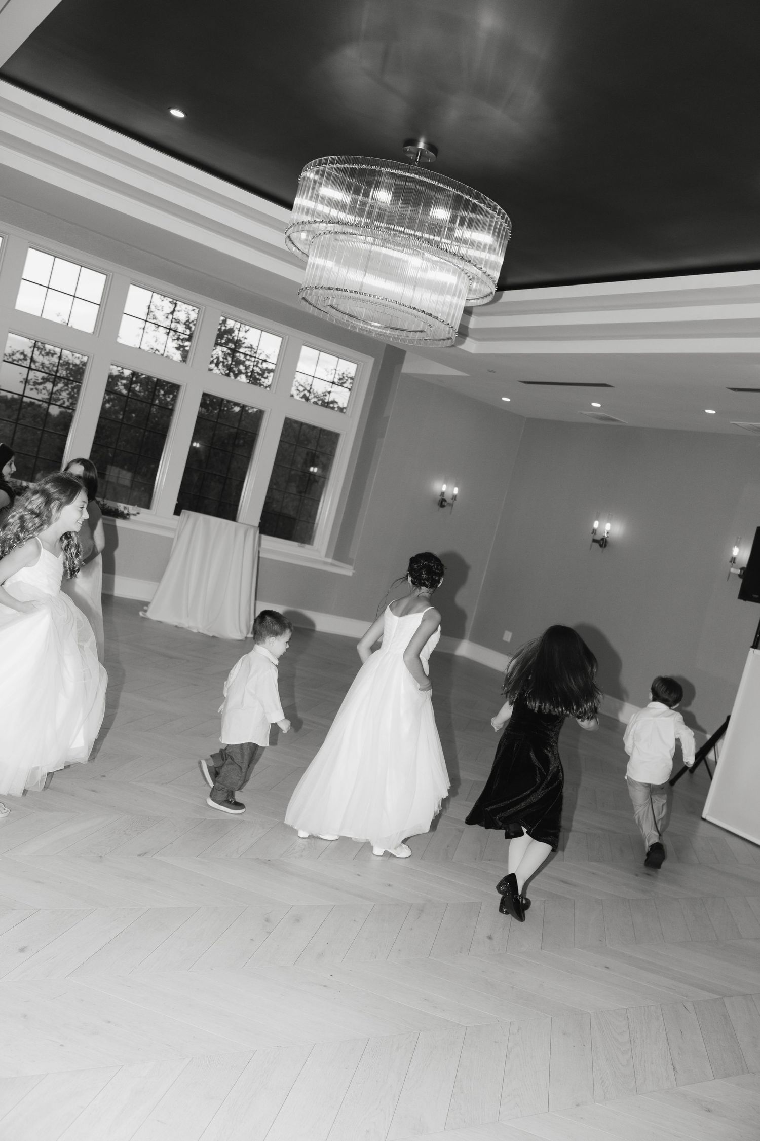 Black and white photo of people dancing in an elegant ballroom with a crystal chandelier and large windows.
