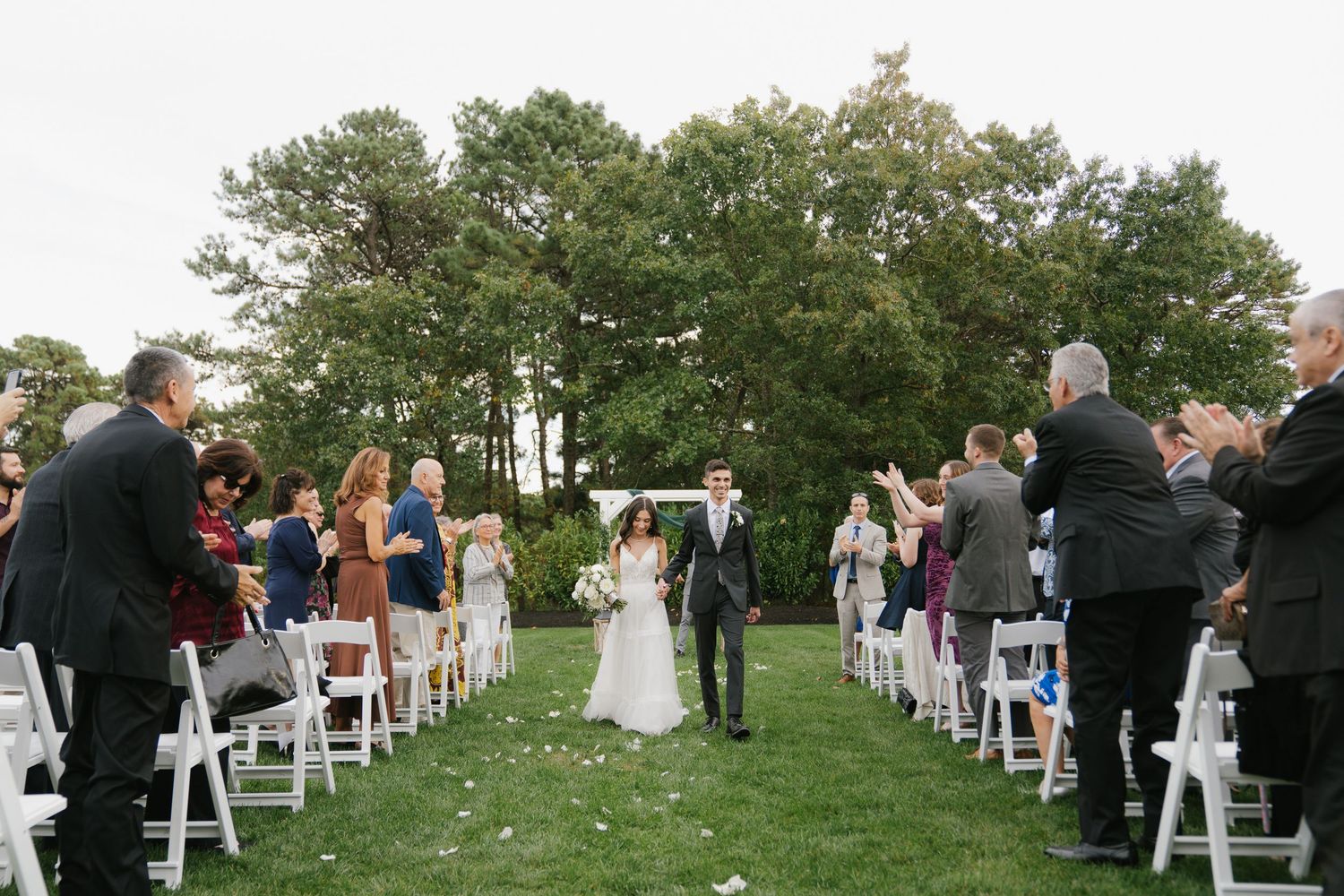 Outdoor wedding ceremony setup with white chairs arranged in rows on a grassy hillside surrounded by trees.