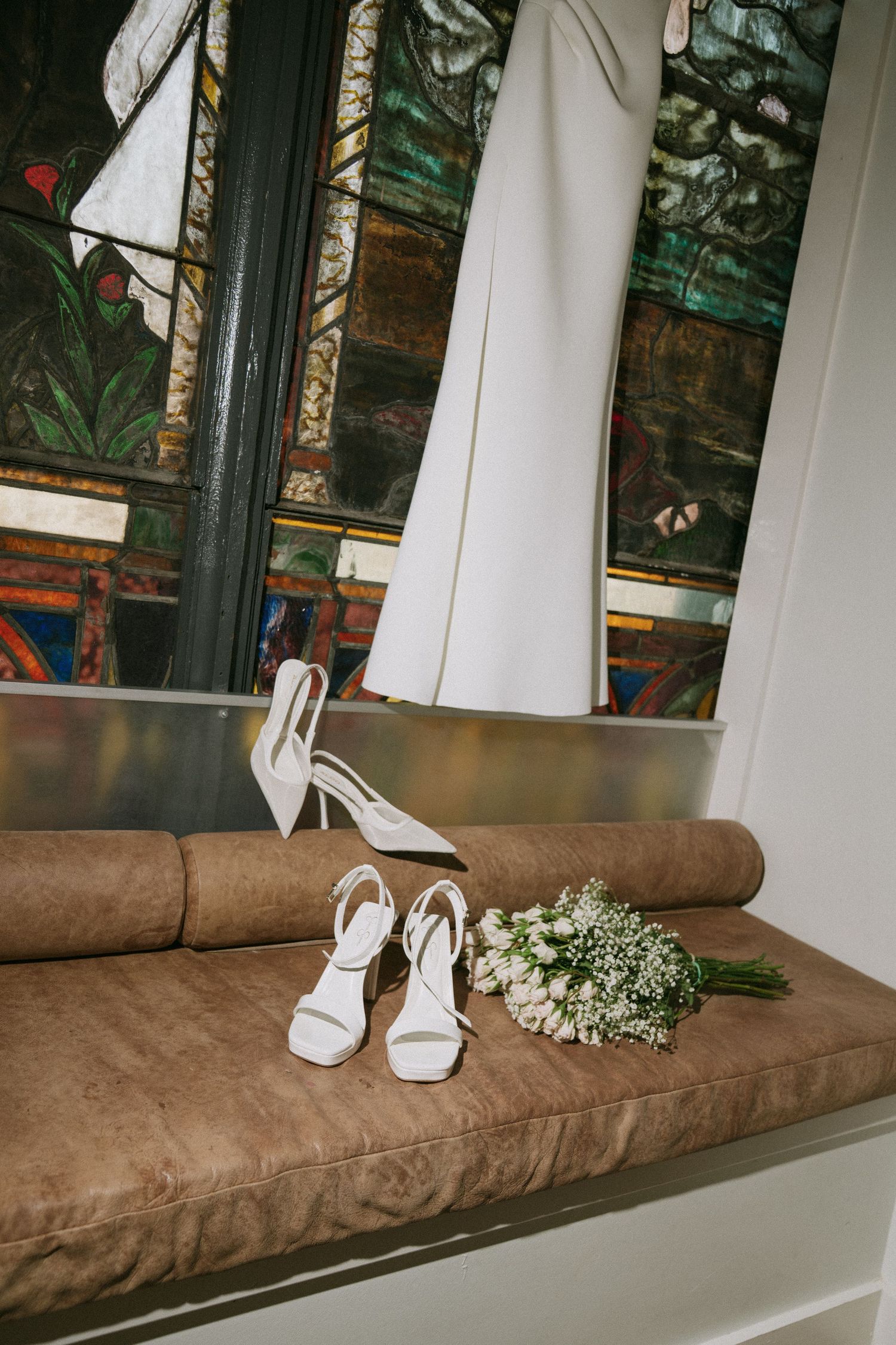 White wedding shoes and baby's breath bouquet arranged on brown bench against stained glass window.