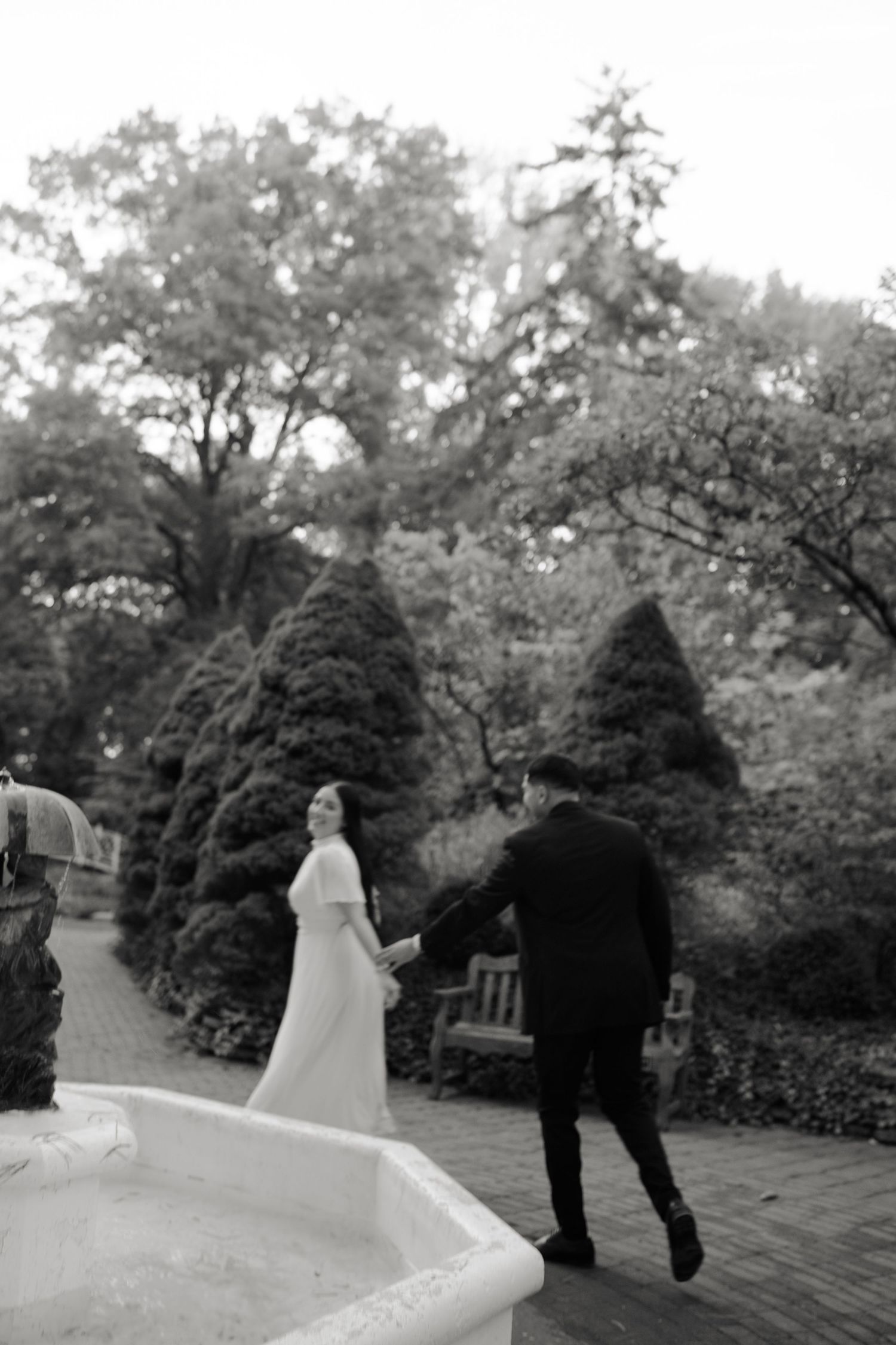 Black and white photo of a bride and groom holding hands near a garden fountain surrounded by evergreen trees.