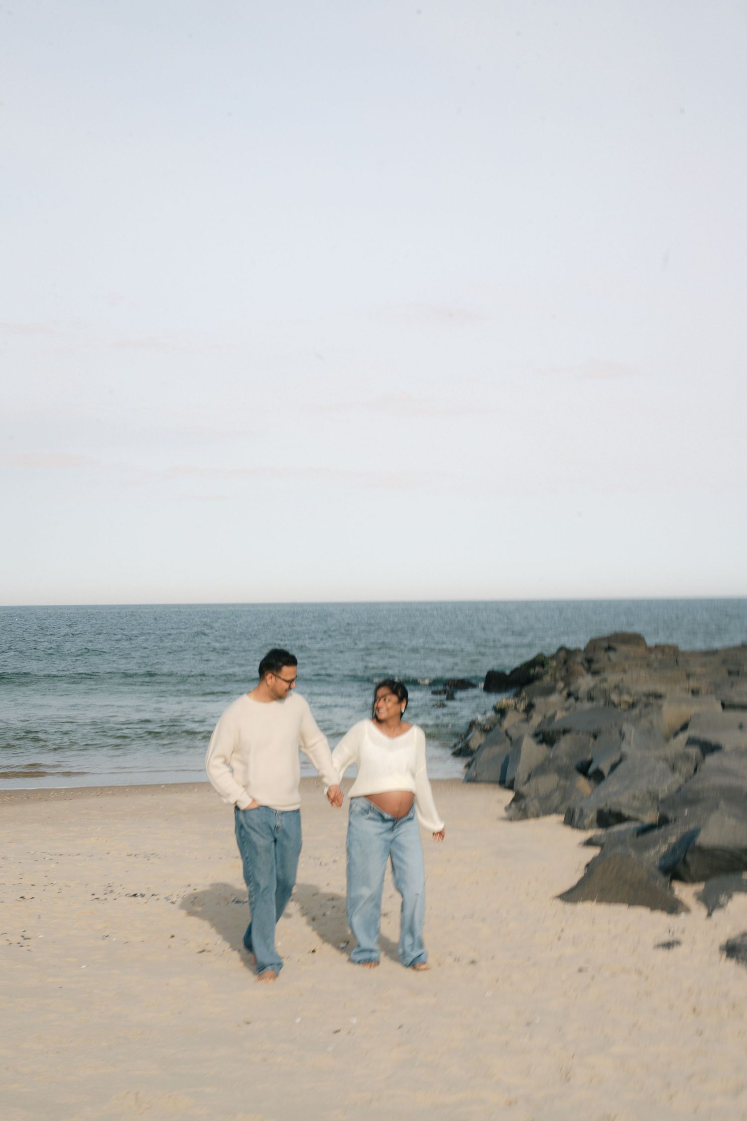 Two people in casual clothing walk together along a sandy beach with rocks and ocean in the background.