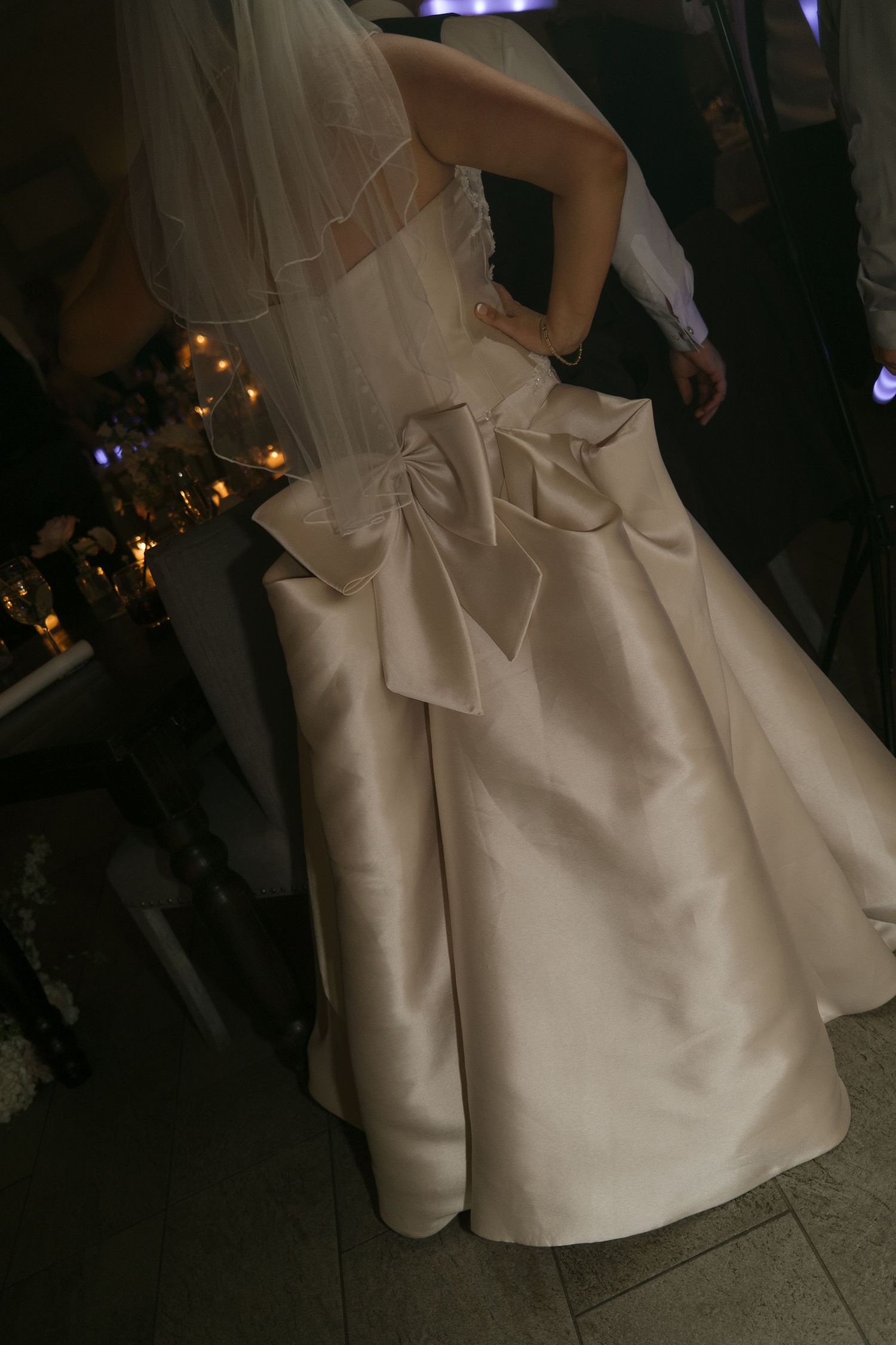 Elegant bride in a white satin wedding gown with large bow detail on the back and flowing skirt at reception.