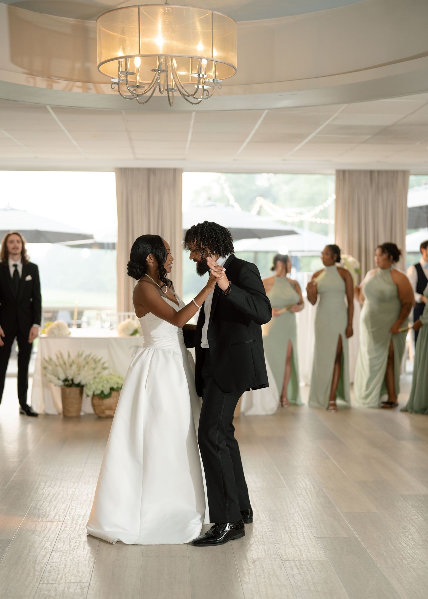 Couple shares romantic first dance under chandelier with guests and bridesmaids in sage green.