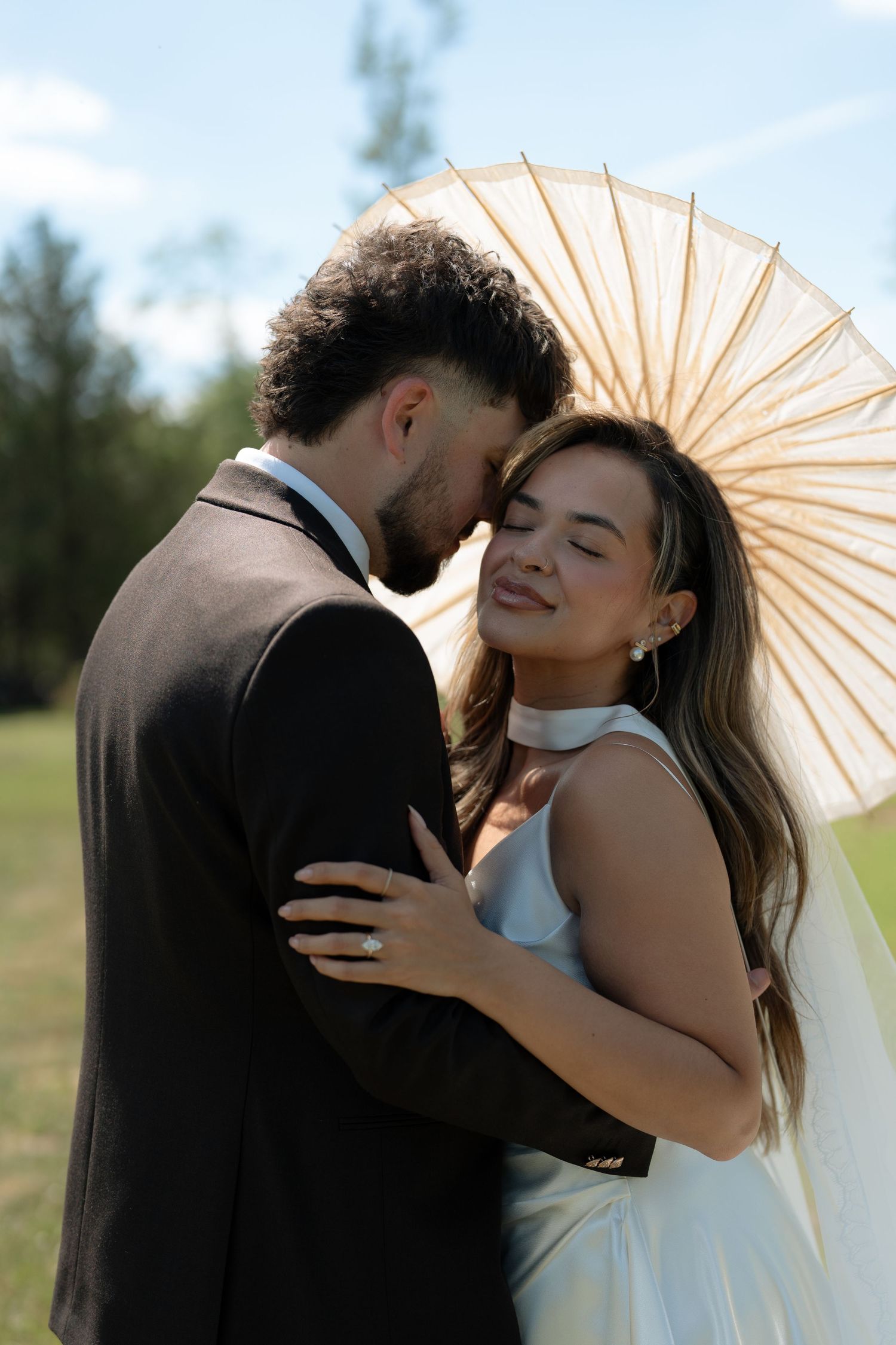 A bride and groom share a romantic outdoor moment, with the bride holding a white parasol umbrella on their wedding day.