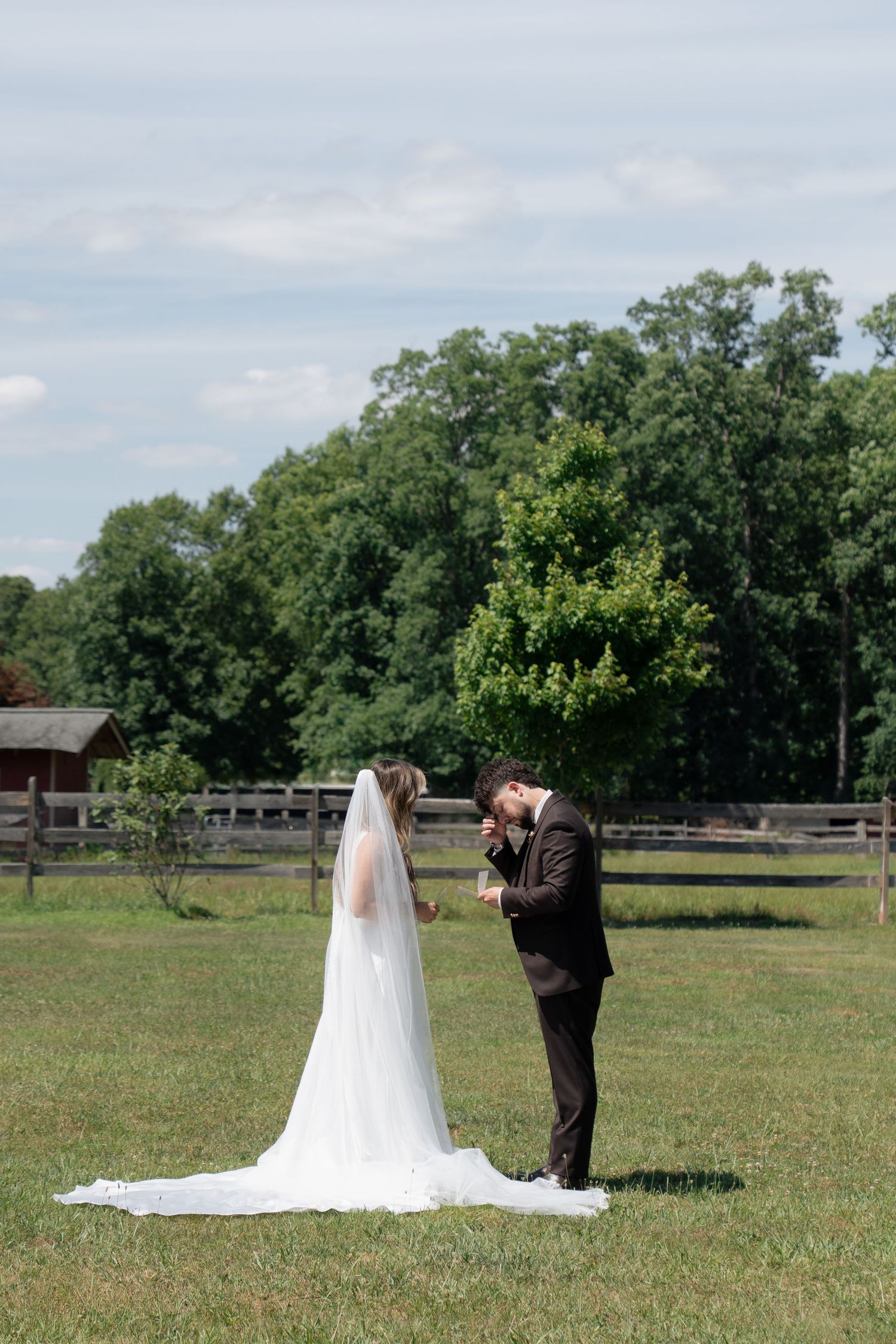 Bride in white gown with cathedral veil and groom holding hands in green farm field.