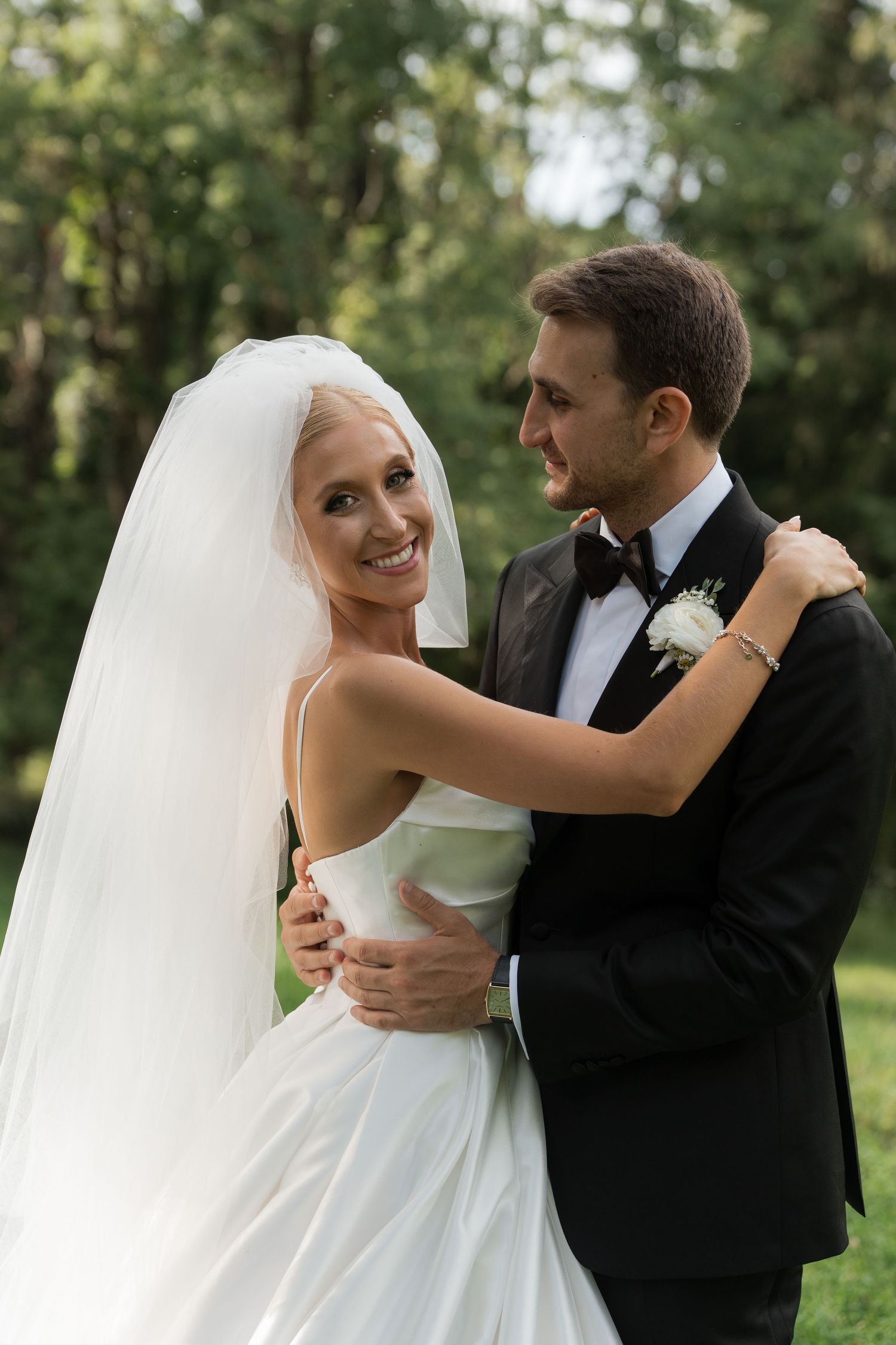 Smiling bride in white gown and veil embraces groom in black tuxedo outdoors at wedding.
