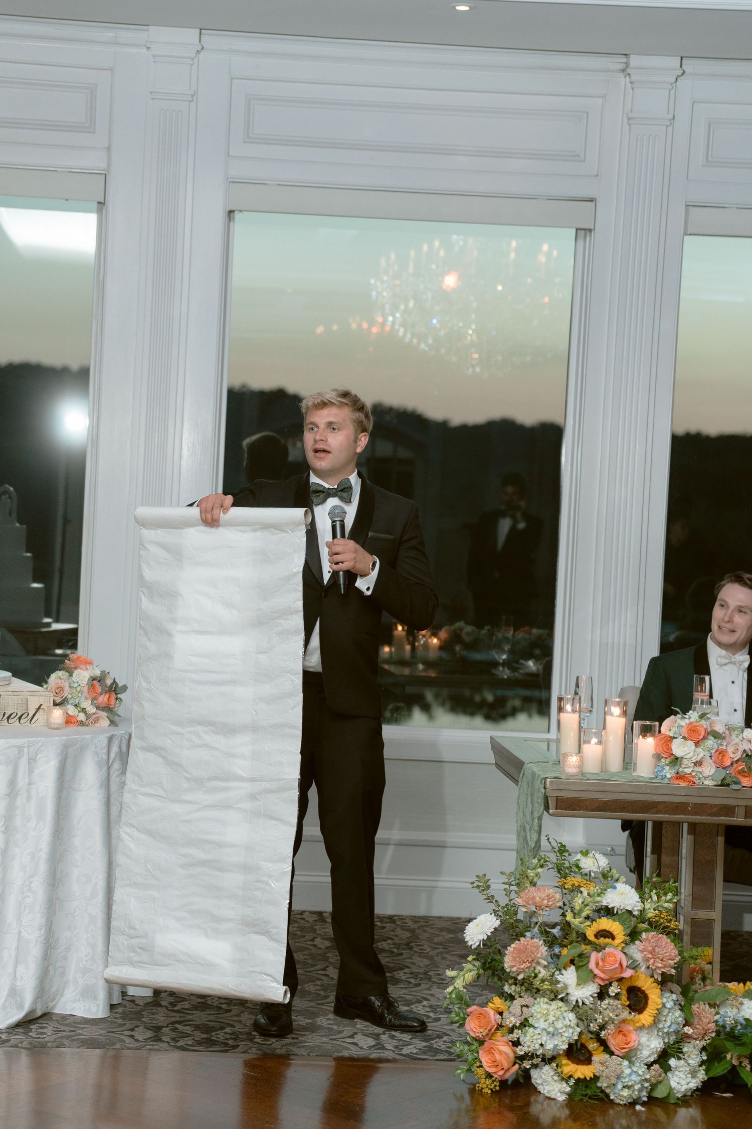 Best man in a tuxedo delivering a heartfelt wedding speech holding a scroll with fireworks visible through the window behind him.