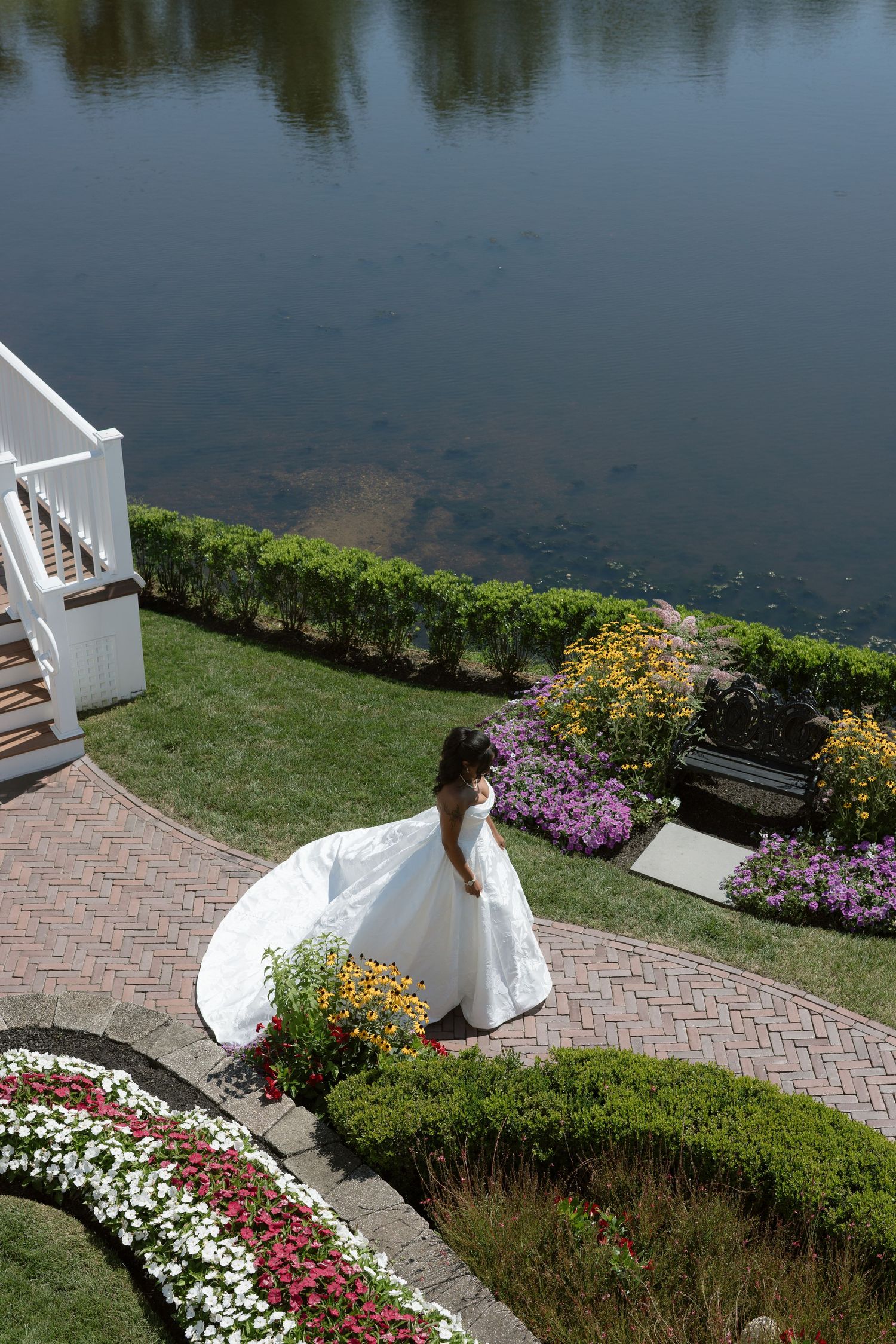 Bride in a flowing white ball gown twirls near a scenic pond surrounded by colorful garden flowers.