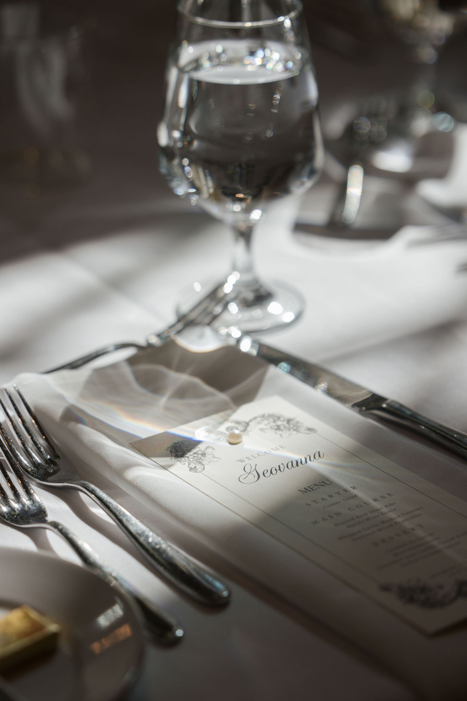 Elegant wedding place setting with personalized menu card reading Giovanna, silver cutlery, and crystal water glass on white linen.