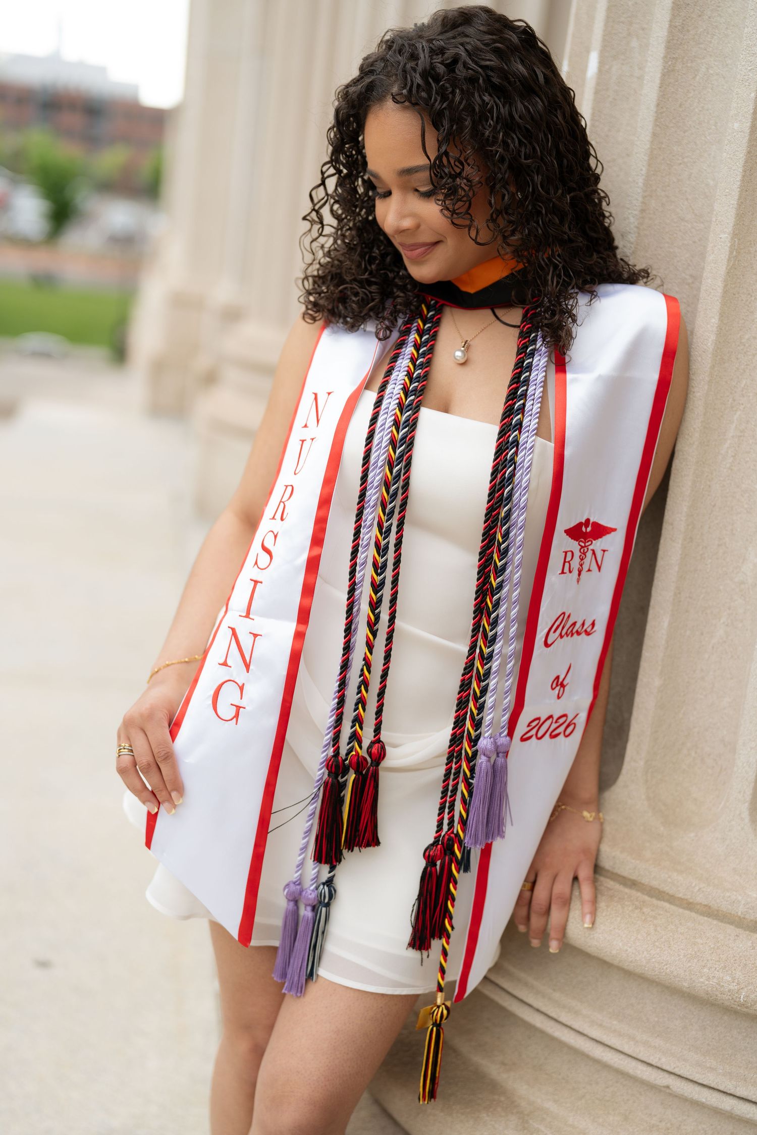 Female nursing graduate leaning on column wearing white dress, nursing stole, and colorful honor cords.