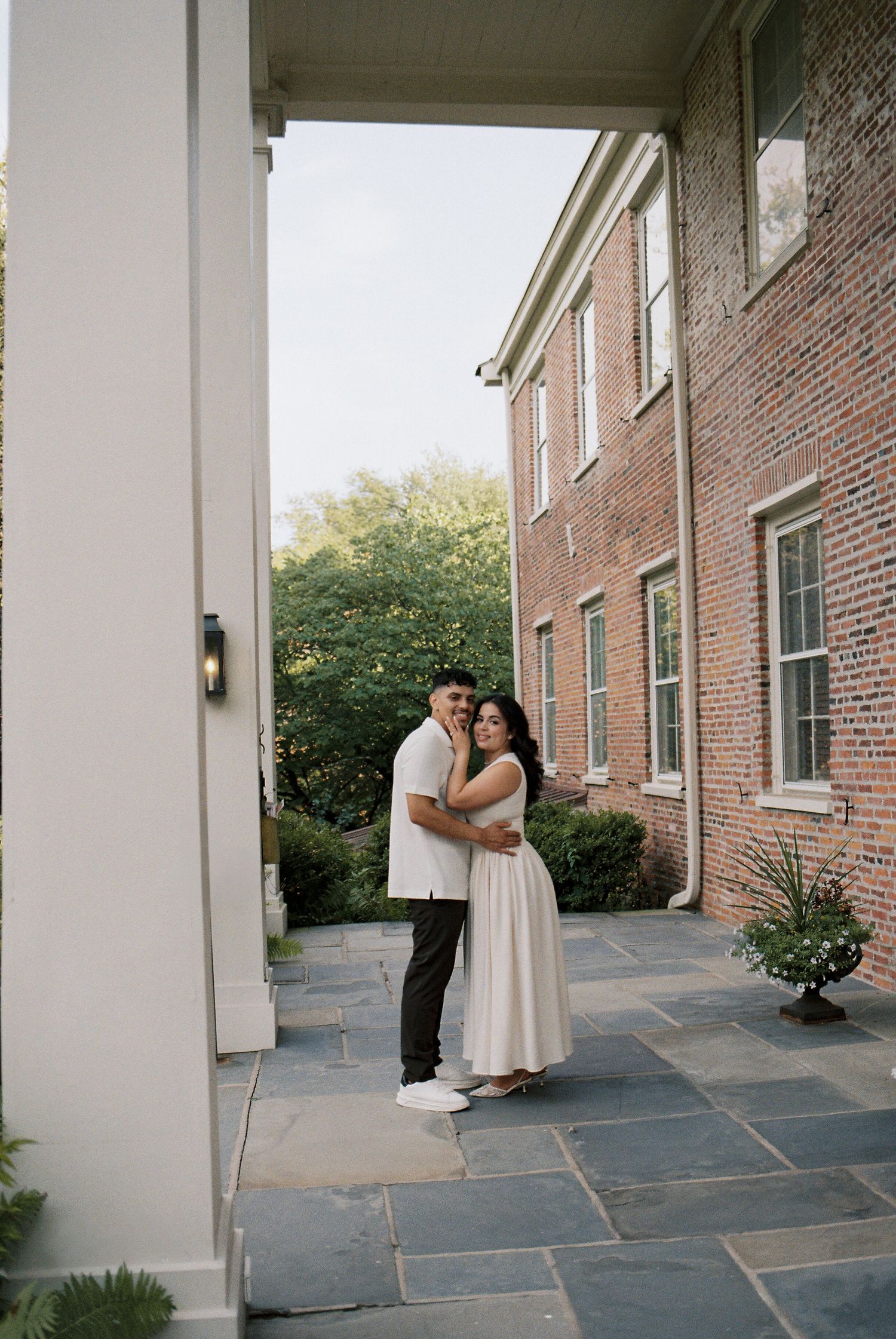 Couple embracing on stone patio beside brick colonial mansion, surrounded by lush greenery.